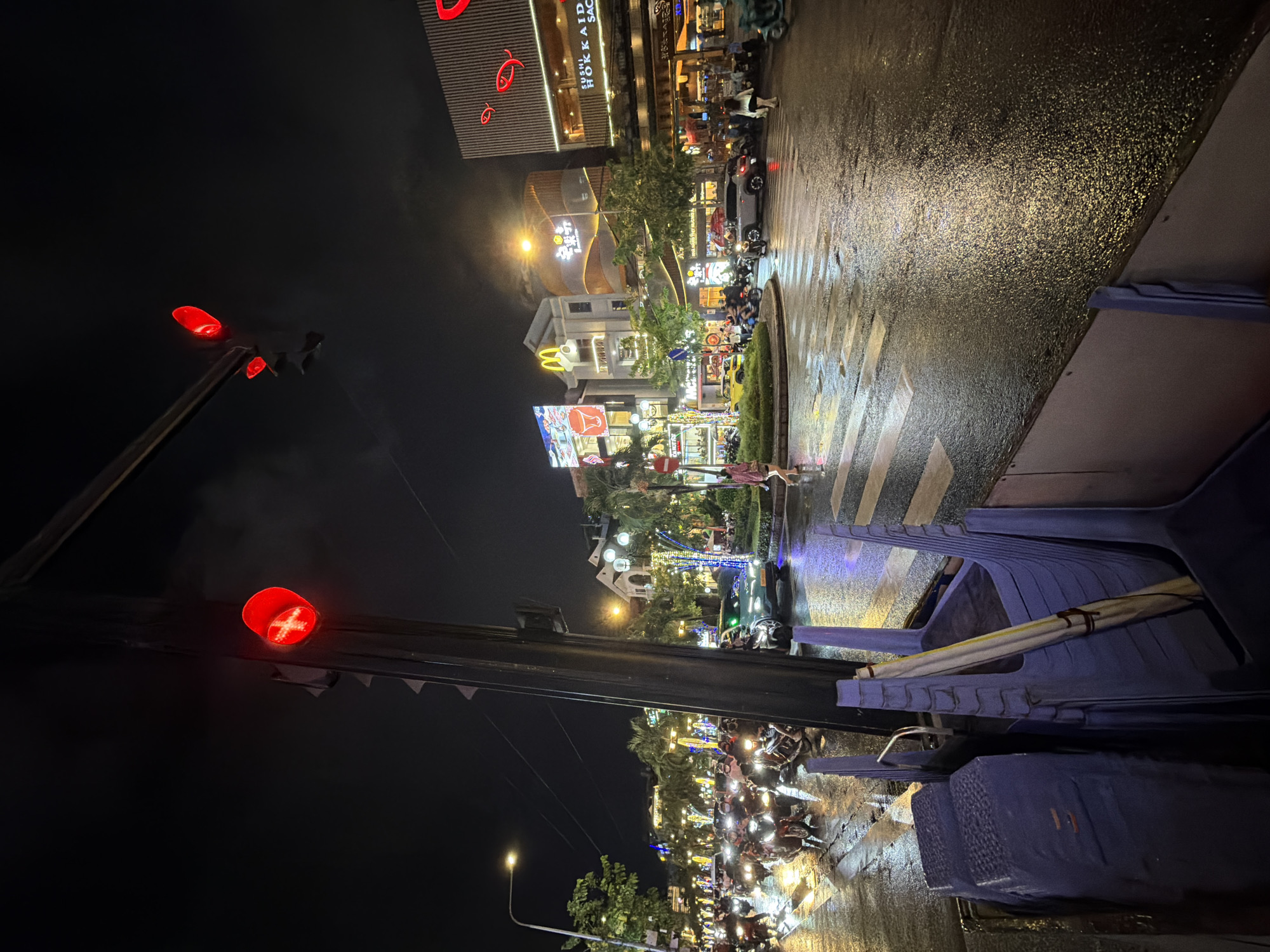 The image depicts a nighttime street scene in Thủ Đức City, Vietnam, likely at an intersection. The road is wet and reflects the surrounding lights, suggesting recent rain. A traffic light is visible in the foreground, displaying a red light with a white cross symbol, indicating a stop. Several motorbikes are stopped at the intersection, indicating traffic. Buildings with illuminated signs are visible on the right side of the street, including one with a "SUSHI HOKKAIDO SACE" sign. On the left side of the street, more buildings with lights and signs, including a McDonald's logo, are present. A few pedestrians are visible on the sidewalks. In the immediate foreground, to the left, are stacked blue plastic chairs, possibly from an outdoor seating area. The overall atmosphere is of a busy urban environment at night, with active traffic despite the red light.