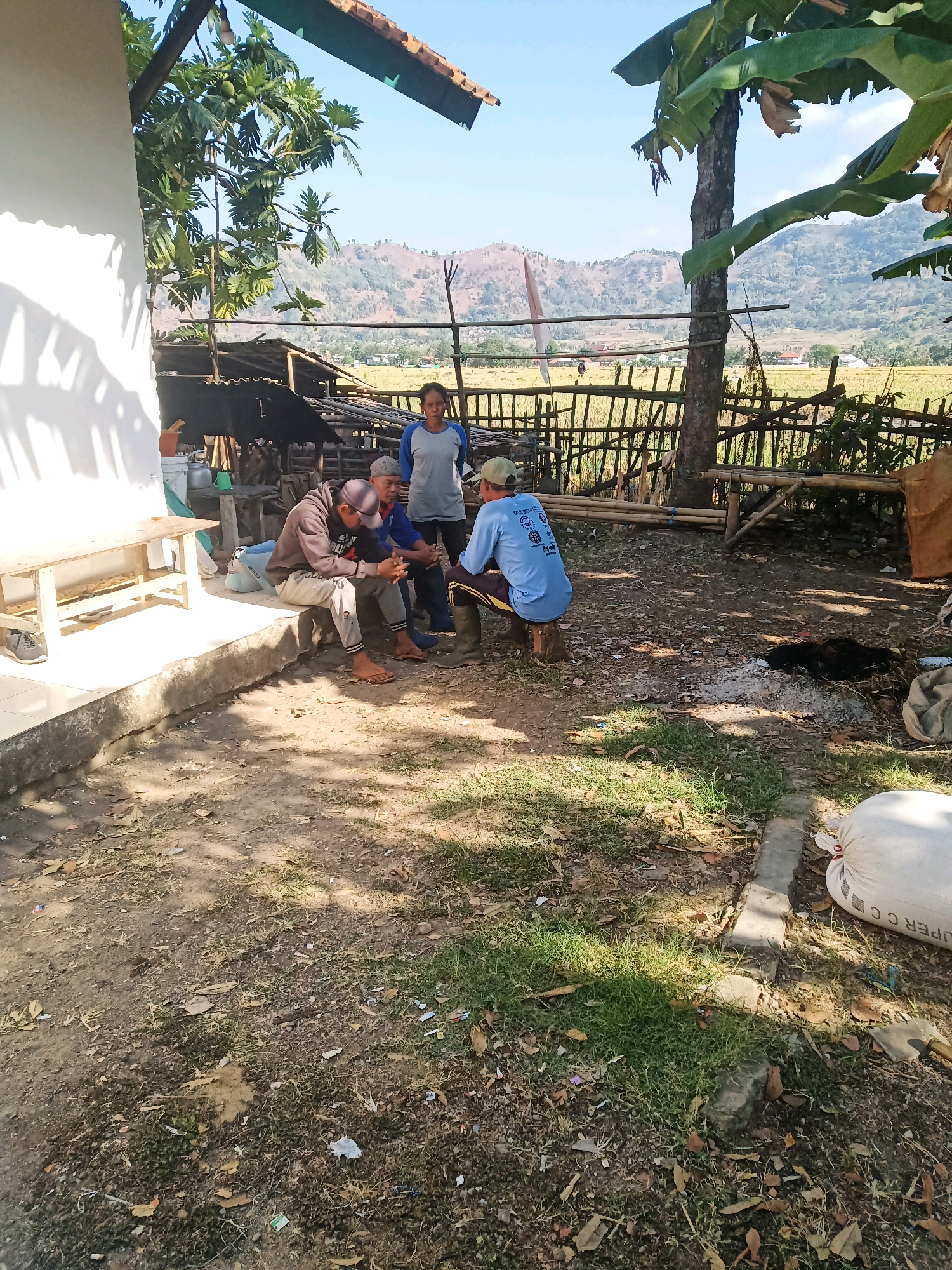 The image depicts an outdoor scene, likely in a rural area, possibly the city of Garut, Indonesia, given the context. Four individuals are present, appearing to be engaged in a conversation. Two men are seated on the ground, one looking at his phone and the other appearing to listen. Another man is seated on a low wooden structure, and a woman stands behind them. The environment is somewhat cluttered, with a wooden bench, bamboo structures, and agricultural fields visible in the background. The ground is a mix of dirt, grass, and fallen leaves. The overall impression is a casual gathering in a rustic setting.
