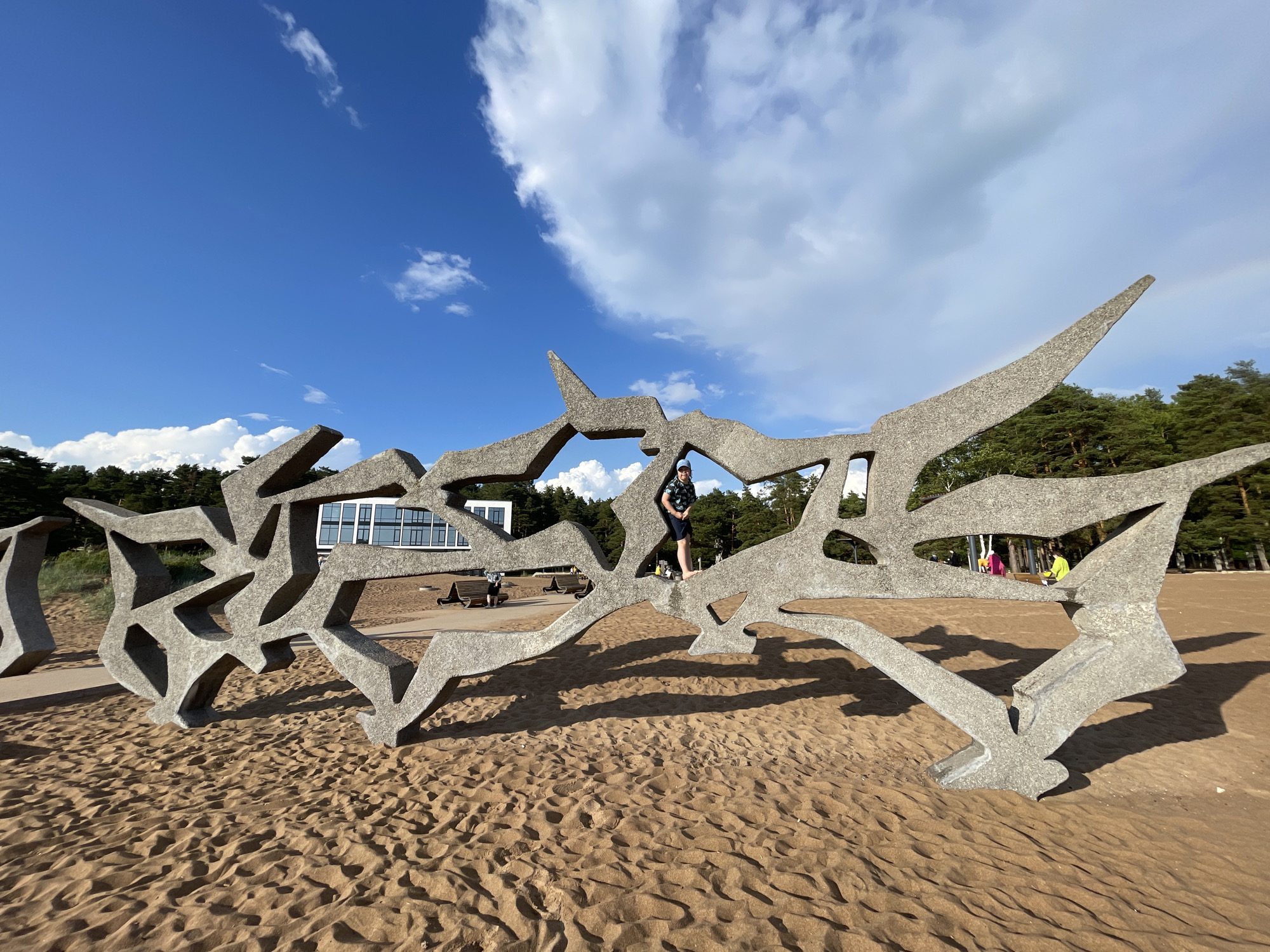 In Solnechnoye, Russia, a person stands on a large, abstract concrete sculpture that resembles a stylized bird or wing. The sculpture is located on a sandy beach, with the rippled sand indicating a dry environment. In the background, a modern building with a glass facade and dark trim is visible, situated behind a line of tall pine trees. Several other people are scattered around the beach, some sitting on benches and others walking. The sky is a bright blue with scattered white clouds, suggesting it is a sunny day, likely in the afternoon due to the long shadows cast by the sculpture. The overall atmosphere appears relaxed and leisurely, consistent with a beach setting on a pleasant day. There is no visible text in the image.
