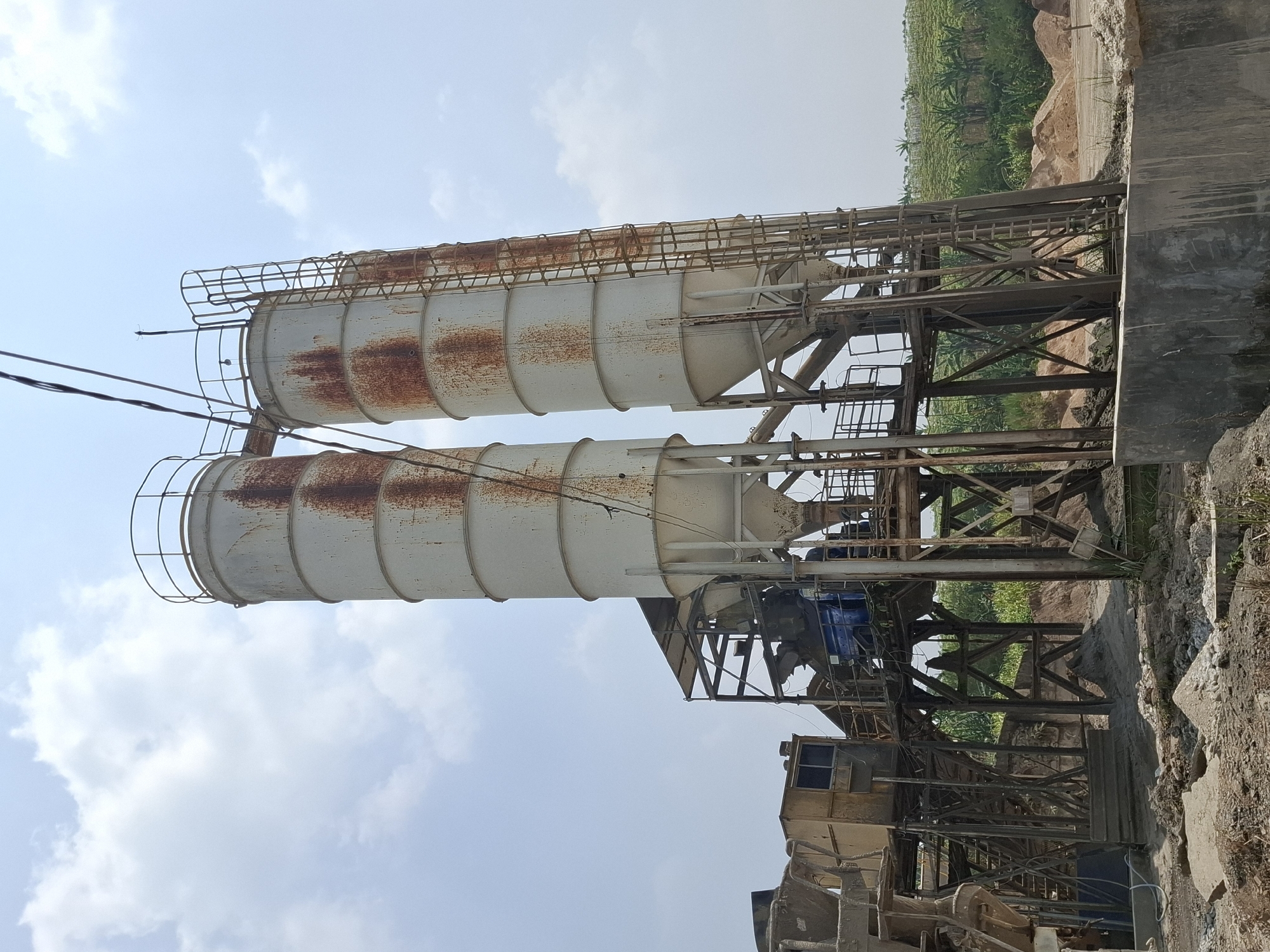 Two tall, white, cylindrical silos, heavily stained with rust, stand side-by-side. Each silo is topped with a railing and has a metal ladder attached to its exterior. They are supported by a complex, multi-level metal framework. Below the silos, a concrete mixing plant with yellow and blue machinery is visible. The ground is covered with dirt, rocks, and piles of sand. In the background, lush green vegetation, likely agricultural fields, stretches towards a hazy horizon under a bright, cloudy sky. The scene depicts an industrial site, possibly a cement production facility, located in Pamuruyan, Indonesia. There are no people visible in the image.