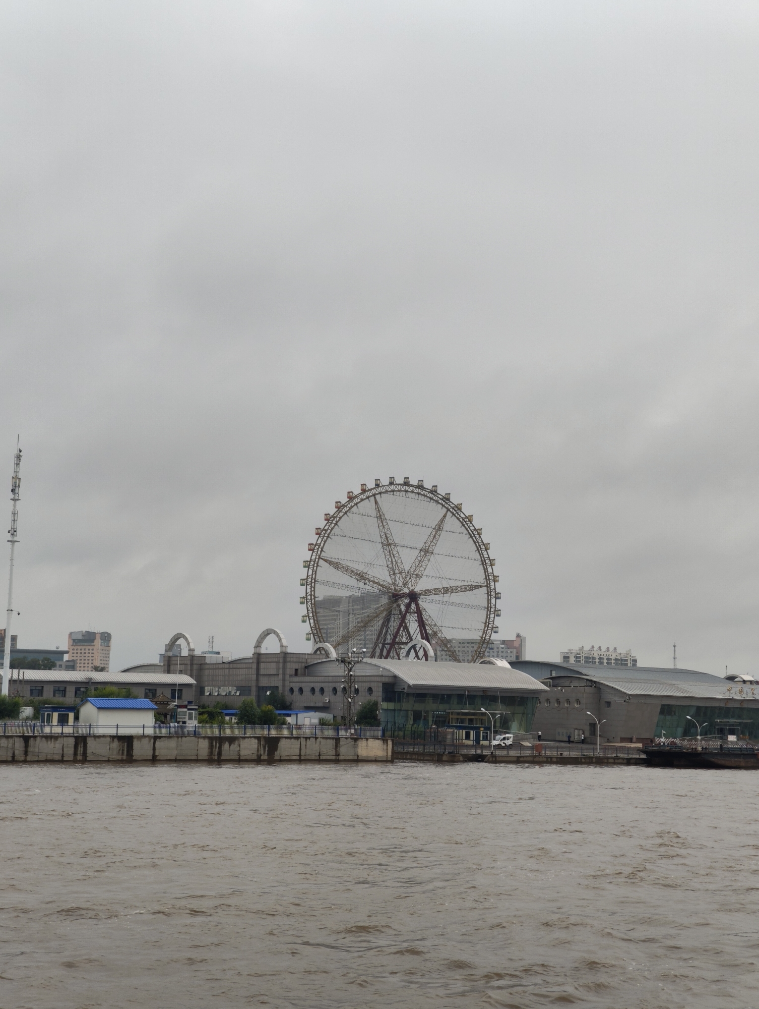 The image depicts a wide, murky river in the foreground, with a concrete embankment forming the middle ground. Beyond the embankment, a complex of buildings is visible, featuring modern architectural elements such as large glass facades and metallic roofing. Dominating the background is a large Ferris wheel, with its distinctive spokes and gondolas. Several other buildings, including what appears to be a tall telecommunications tower on the left and a cluster of high-rise structures in the distance, contribute to the urban landscape. The sky is overcast, casting a dull light over the scene. The environment suggests an urban waterfront, likely in 幸福乡, China, given the provided context. There are a few scattered individuals and vehicles visible on the embankment and near the buildings, but no specific interactions are discernible.
