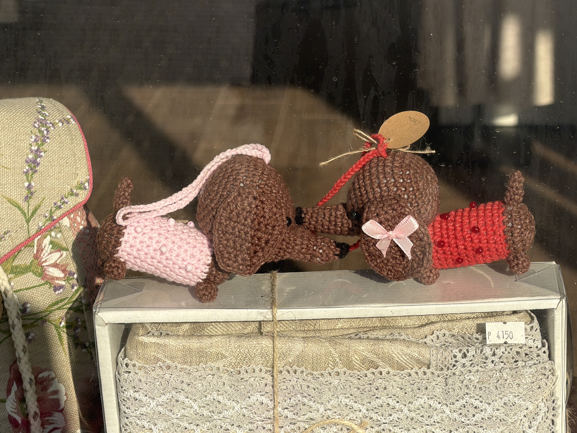 In Vologda, Russia, a display window showcases three crocheted dachshund figurines. The figurines are brown, with one wearing a pink knitted sweater and the other a red knitted sweater adorned with small red beads. A pink bow is attached to the red-sweatered dog's head. The dachshunds are arranged on a white box containing rolled fabric, one brown and one light beige, both textured with patterns and edged with white lace. The background is a dark, reflective surface, possibly a window or glass door, showing distorted reflections of vertical white elements, suggesting indoor lighting or blinds. To the left, a fabric item with a floral pattern, featuring lavender and pink flowers on a beige background, is partially visible, with a rope handle. A price tag with "P 4150" is attached to the fabric display.