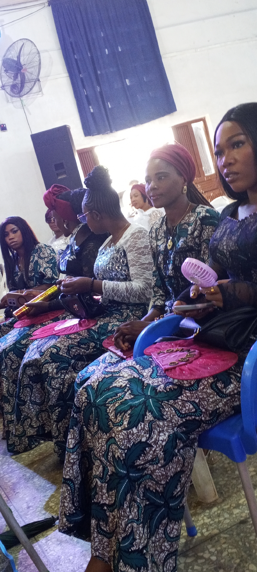 The image shows a row of seated women indoors, likely at an event. The women are dressed in similar patterned clothing, possibly a uniform for the occasion. Some hold handheld fans. In the background, there's a wall-mounted fan, a speaker, and curtains. The location appears to be Sabon Tasha, Nigeria.