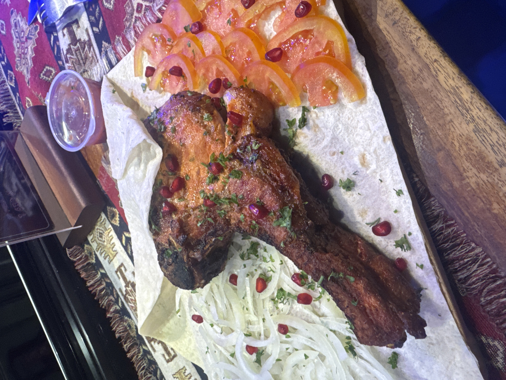 The image shows a dish of grilled meat, likely lamb or mutton ribs, served on a flatbread with sliced tomatoes and onions, garnished with pomegranate seeds and herbs. A small plastic container sits nearby. The scene is set on a table covered with a patterned cloth, potentially in North Kuta, Indonesia.