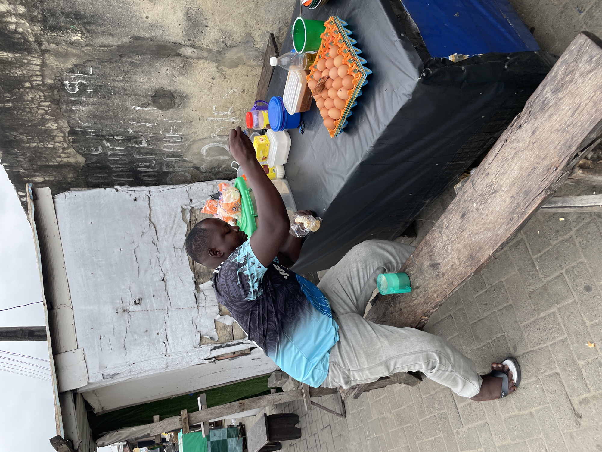 A Nigerian man is pictured preparing food at a street stall in Ado, Nigeria. The stall is made of corrugated metal and weathered wood, with a black tarp covering the table. He is wearing a blue and black patterned t-shirt and grey pants, with a flip-flop on his left foot. In front of him are several items for sale, including a carton of eggs, bottles of oil, and containers of food. He is holding a small piece of bread and reaching for an egg. The background shows a graffiti-covered wall and more makeshift stalls. The scene appears to be an outdoor market on a cloudy day.