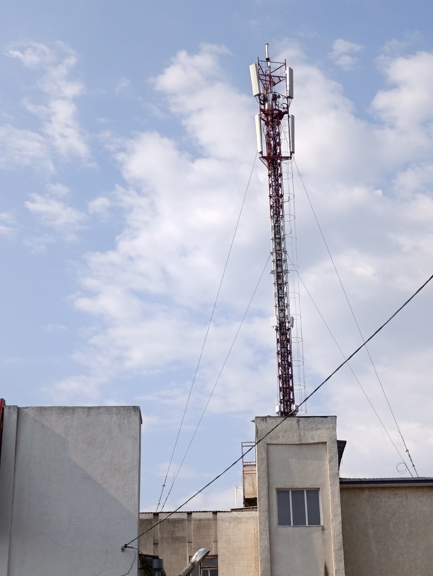 A tall, red telecommunications tower stands prominently against a pale blue sky dotted with white clouds. The tower is constructed of a lattice of metal beams and features multiple antennas and equipment attached to its structure. It is anchored by several guy wires, some of which extend to the rooftops of adjacent buildings.
In the foreground, parts of two buildings are visible. The building on the left is a light-colored, flat-roofed structure with a large, blank wall. The building on the right is a more complex structure, also light-colored, with a prominent vertical section featuring a small, rectangular window with three panes. This section appears to be a part of a larger building that extends out of view. The overall scene depicts an urban or semi-urban environment, likely in the town council area of Dubăsari, Moldova, given the context provided. There are no people visible in the image.