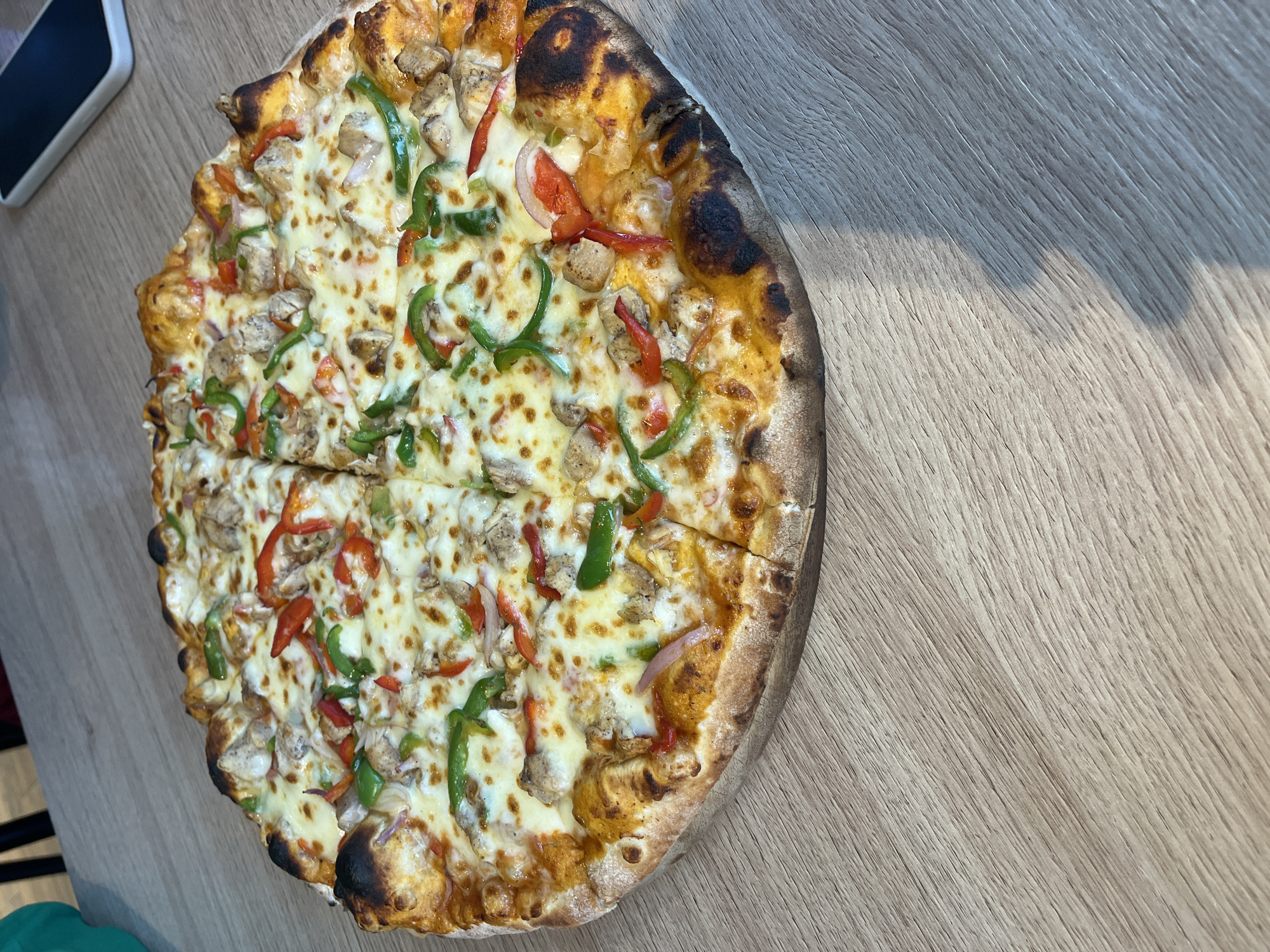 An overhead, close-up photograph displays a large, round pizza, segmented into eight slices, situated on a light-colored wooden table. The pizza exhibits a thick, well-baked crust with visible charring at its edges. Toppings include a uniform layer of melted cheese, diced chicken pieces, green bell pepper strips, red bell pepper strips, and thinly sliced red onion. The wooden table surface features a pronounced diagonal grain pattern. A significant shadow is cast on the lower right portion of the table. In the upper right, a white smartphone rests face down. A partial view of a teal object and dark, indistinct shapes are visible in the upper left corner. No individuals are present, and no active interactions are depicted. This scene is situated in Abuja, Nigeria.