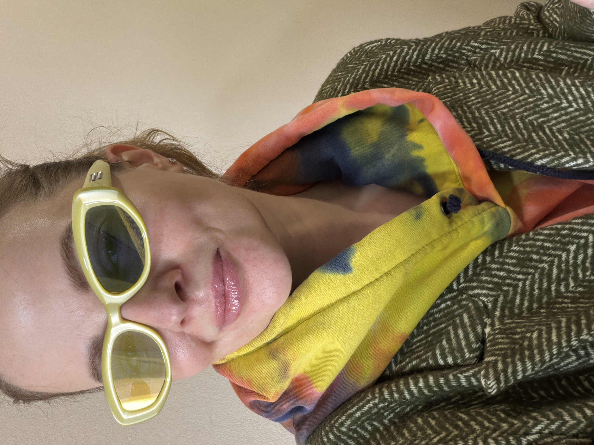 A woman is photographed up close, likely indoors given the even lighting and wall in the background. She is wearing pale yellow, oval-shaped sunglasses. She is also wearing a hoodie with a colorful tie-dye pattern, which is beneath an olive-green tweed jacket. There is no indication of the location being Moscow, Russia.