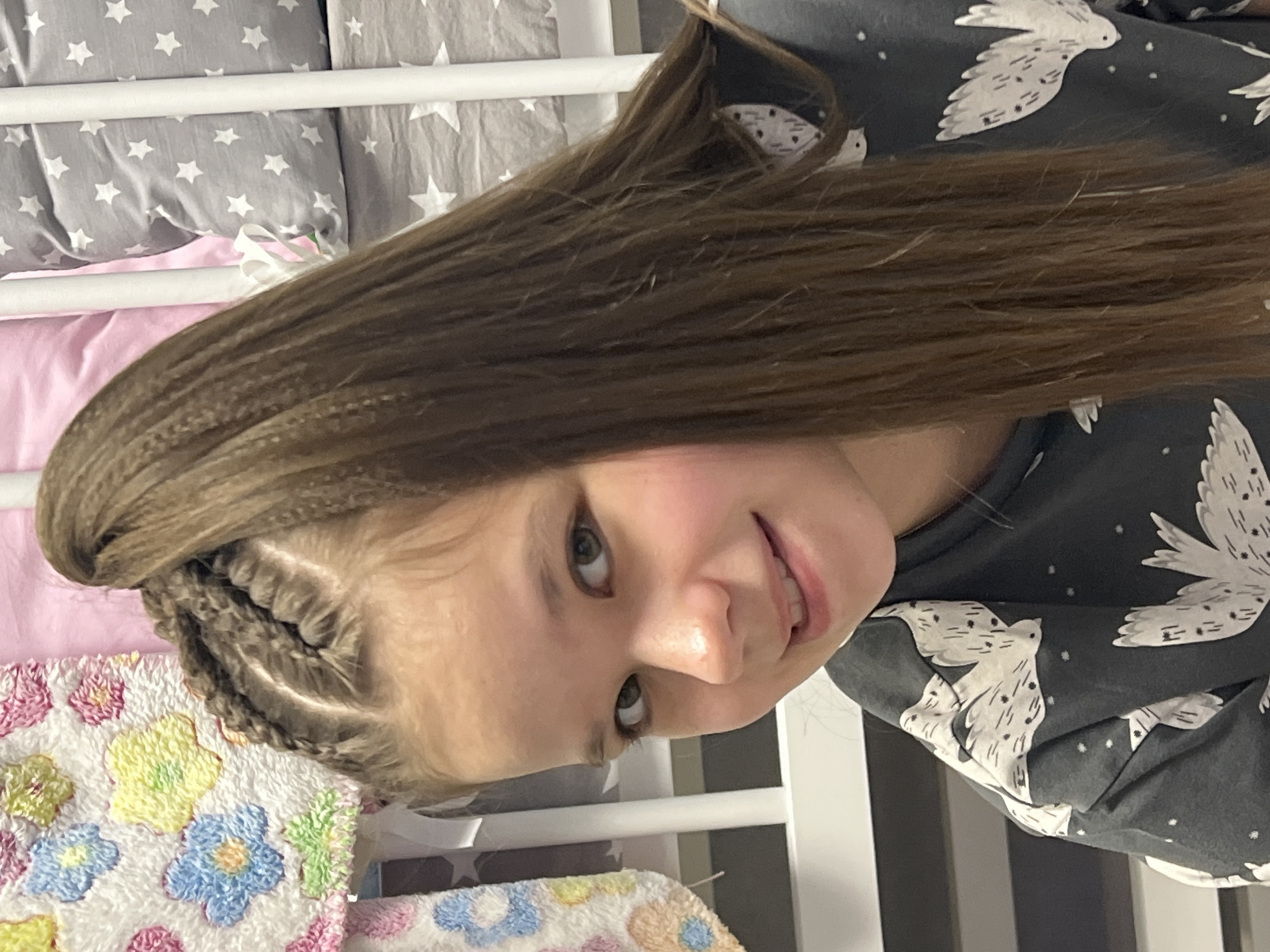 A young girl is pictured indoors, likely in a bedroom. She has brown hair styled with braids along her hairline that transition into crimped waves. She is wearing a grey top with a white bird print. In the background, there is a baby cot with grey star-patterned pillows and a pink sheet. There is also a patterned blanket with floral designs. Location is not discernible from the provided image.