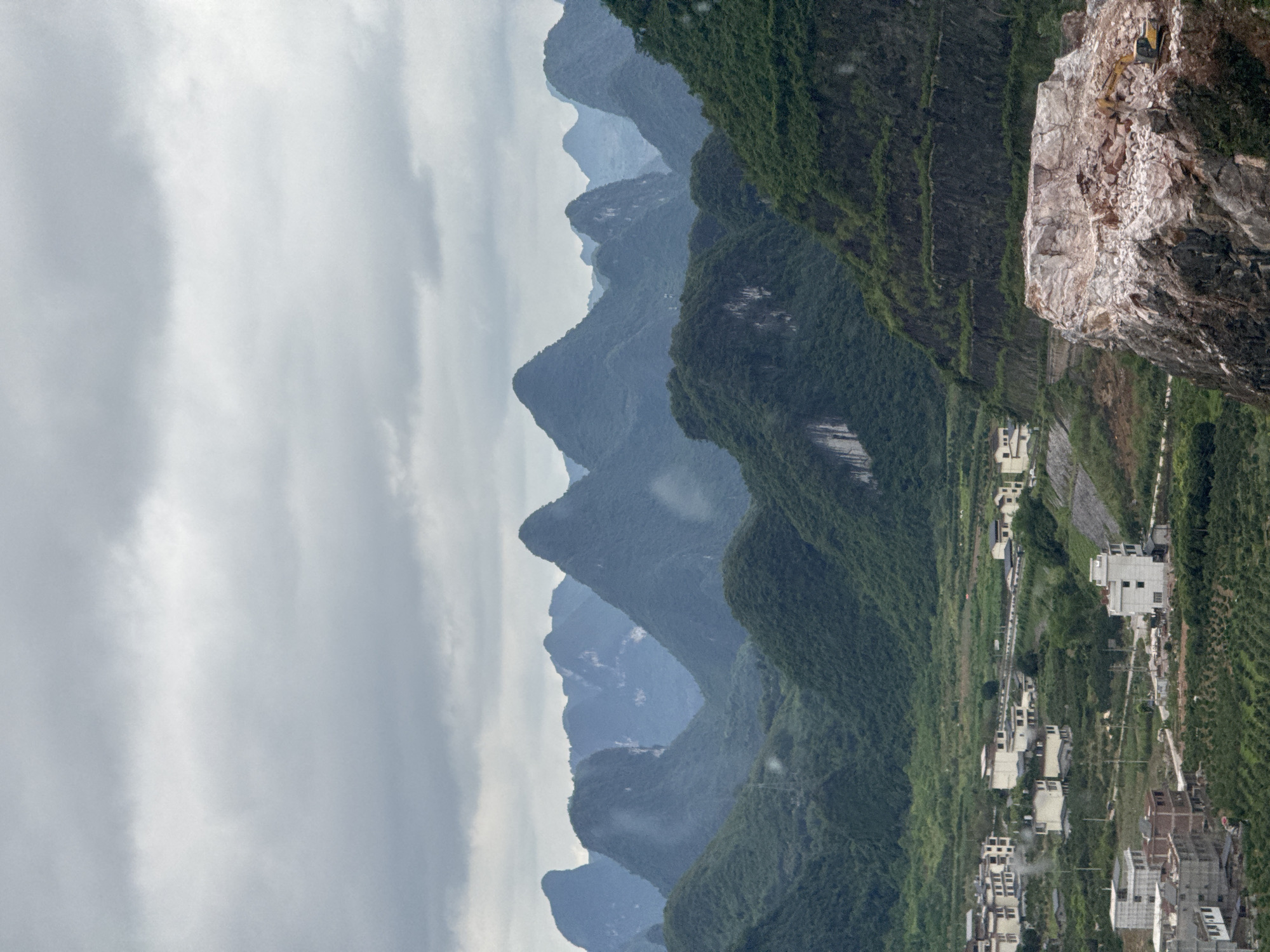 This landscape photograph shows the city of Guilin, China, in an outdoor setting featuring a towering rock feature to the right. The city features a series of buildings with predominantly white facades and dark-colored roofs. A paved road runs parallel to the buildings. The background shows a series of mountains, some of which are covered in green vegetation, fading into the distance under an overcast sky. Atop the towering rock to the right, there appears to be construction equipment. The image is oriented vertically.