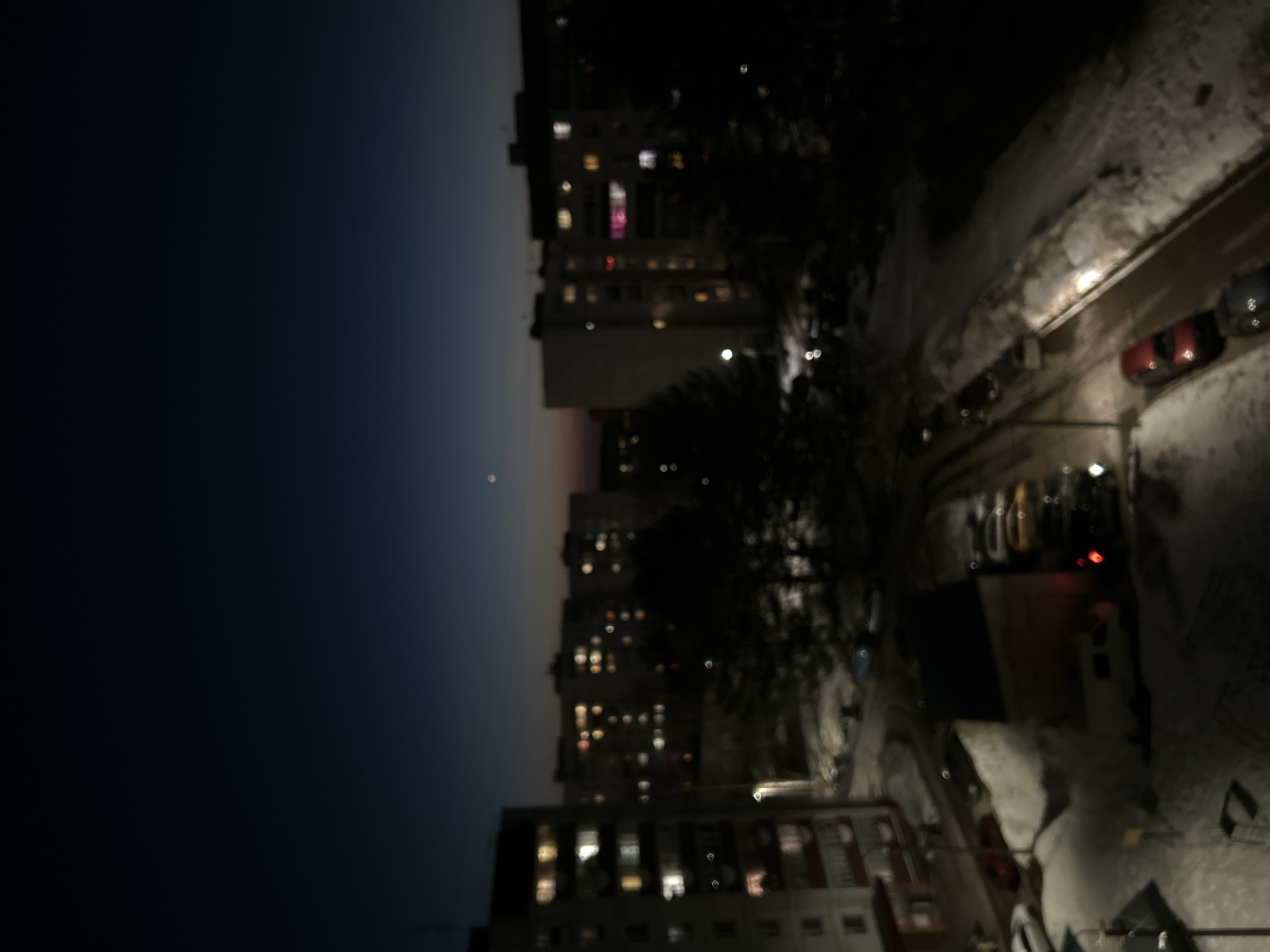 The image depicts a night scene in what appears to be a residential area of a city. Tall apartment buildings with lit windows rise into a dark sky, with a faint glow on the horizon. The street level shows parked cars along a snow-covered street illuminated by streetlights. There are trees between the buildings and the street. The blur of the image obscures details of people and interactions. Given the prompt, this is likely a nighttime view in Chaikovsky, Russia, although without further evidence, the precise location cannot be determined.