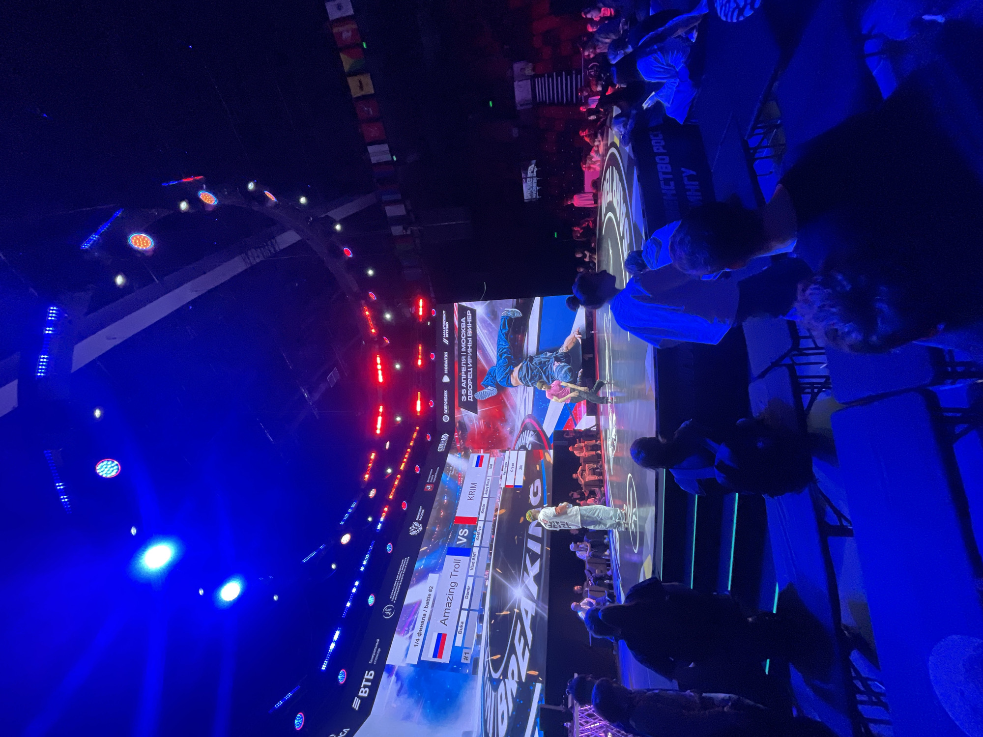 The image depicts a breakdancing competition, likely a quarterfinals battle between "Amazing Troll" and "KRIM". The event takes place in Moscow, Russia, from April 3-5, at the Irina Viner Palace. The scene is illuminated with blue lighting, and a large screen displays the competitors, their names, and judges. Some spectators are visible, watching from their seats. Sponsorship logos such as Gazprombank, Novatek, and VTB are also present on the screen.