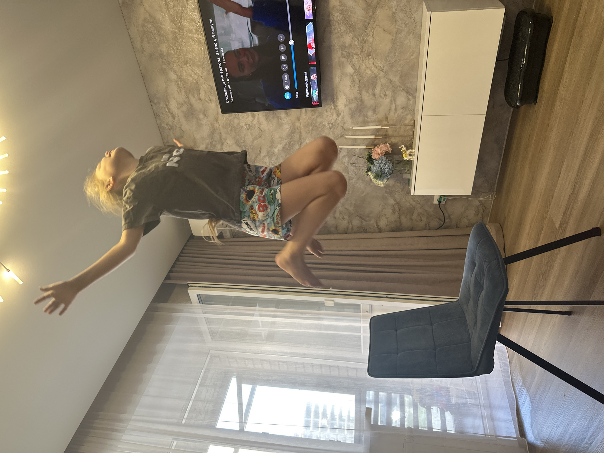 A young person with blonde hair is jumping in a living room. They are wearing a dark grey t-shirt and colorful shorts with a graphic print. Their arms are outstretched upwards, and their head is tilted back, with a joyful expression. The room has a modern aesthetic with a textured grey wall, a large television mounted on the wall displaying a TV show, and a white floating cabinet below it. A dark blue chair with a modern design is positioned to the left of the frame. A window with sheer curtains is visible on the far left, allowing natural light to enter the room. The floor is made of light-colored wood. The scene suggests a moment of spontaneous activity and enjoyment within a domestic setting.
