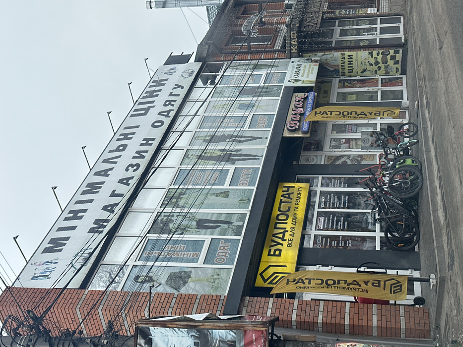The image shows the exterior of a storefront in Tarasivka, Ukraine, located at coordinates 50.343, 30.303. The storefront is made of brick and glass and features a large sign reading "MINIMALNI TSINI MAGAZIN ODYAGU" in Ukrainian, which translates to "Minimal Prices Clothing Store". There are also two smaller signs, one for "BUDPOSTACH" and one for "VAUAK". The BUDPOSTACH sign reads "VSE DLYA DOMU TA REMONTU", which means "Everything for Home and Repair" in Ukrainian. The VAUAK sign reads "KOSHETKA KURSOVA DZ VYRROBNITSA" in Ukrainian, which means "The Best Coursework for Production". The storefront has several large windows displaying clothing and shoes. The storefront is flanked by two yellow flags with the logo of a company called "NATSOZAPOLYAVB". Four bicycles are parked outside the storefront. The street is paved and appears to be relatively quiet with few pedestrians visible. The sky is overcast.