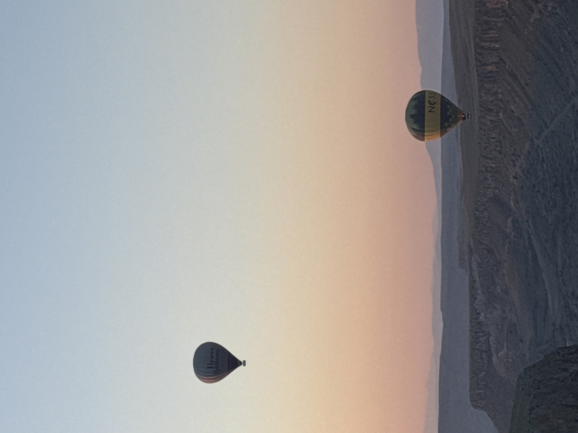 The image captures an aerial view of two hot air balloons drifting peacefully above a dramatic, rugged landscape, likely during sunrise or sunset. The sky displays a beautiful gradient, transitioning from a pale blue at the top to soft peach and orange tones closer to the horizon. Below the sky, an expansive terrain unfolds, characterized by deep valleys, steep cliffs, and distinctive geological formations typical of the Cappadocia region in Yeşilhisar, Turkey.
In the mid-ground, a larger hot air balloon is prominent, featuring a pattern of dark green and yellow stripes. The company name "CAPPADOCIANESS" is clearly visible on its side. Further in the distance, a second, smaller hot air balloon is seen, appearing dark brown or black, with the text "Heves Balloons" inscribed on its surface. Both balloons are airborne, indicating a hot air ballooning event or tour. The weather is clear and calm, offering excellent visibility of the expansive scenery. While no people are explicitly visible, their presence is implied by the operation of the balloons.