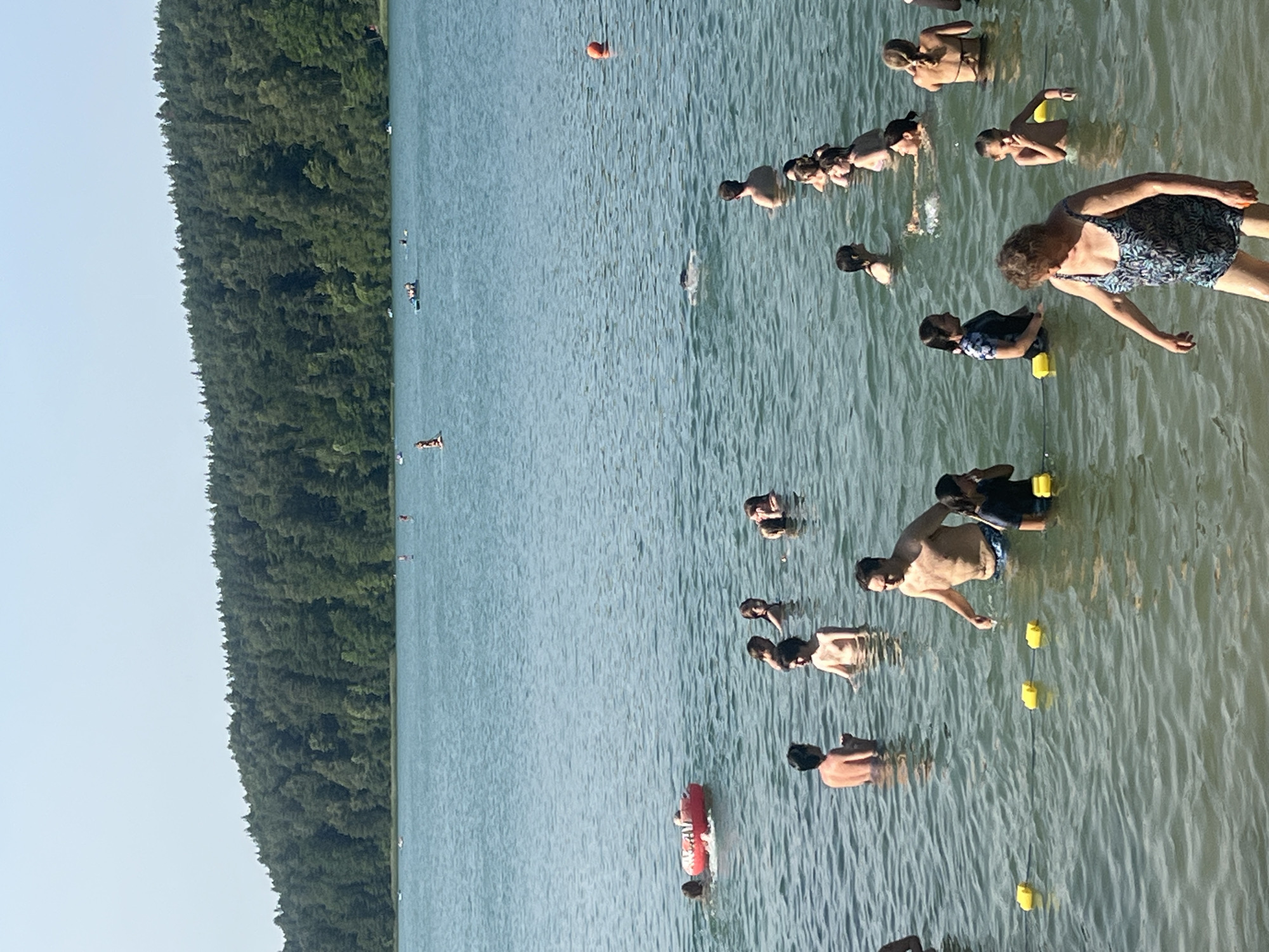 The image depicts a scene at a body of water, likely a lake or a large pond, with a wooded shoreline in the background. Several people are visible in the water, engaged in various activities such as swimming and wading. In the foreground, an older woman in a patterned swimsuit is walking in the shallow water, looking down. Further back, a man in swimming trunks is standing in the water, gesturing with his hand. A group of people, including children, are clustered together in the middle ground. In the background, other individuals are scattered across the water, some on what appear to be small boats or paddleboards. The water is calm with small ripples. The sky is clear and light. The overall impression is a recreational gathering at an outdoor aquatic location. Based on the context of Vilnius, Lithuania, this scene is likely from one of the city's popular swimming areas, such as the beaches of the various lakes surrounding Vilnius, for example, the beaches of Tapeliai lakes or the shores of Lake Balsys.
