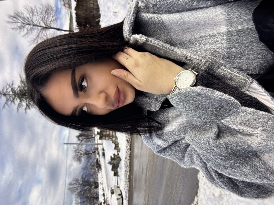 An outdoor, eye-level, slightly off-center portrait features a female individual against a snow-covered landscape in the city of Zhabokrat, Bulgaria. The subject, positioned in the right foreground, has long, dark, straight hair, prominent eyebrows, and full lips, looking upwards and to her right. Her right hand is raised, with fingers resting near her chin; a small, dark tattoo (possibly "A.C.J.") is visible on the lateral aspect of her right index finger. She wears a silver-toned watch with a round face and beaded strap on her left wrist. Her attire includes a grey, textured, possibly plaid, collared jacket over a dark garment.
The background depicts a winter scene with snow covering the ground, including a partially visible asphalt road to the left. Several bare deciduous trees and some evergreen foliage are present. A light-colored vehicle is parked on the snowy road shoulder to the far left. A low stone wall lines the road. Distant, snow-dusted hills or mountains are visible under an overcast, light grey sky.