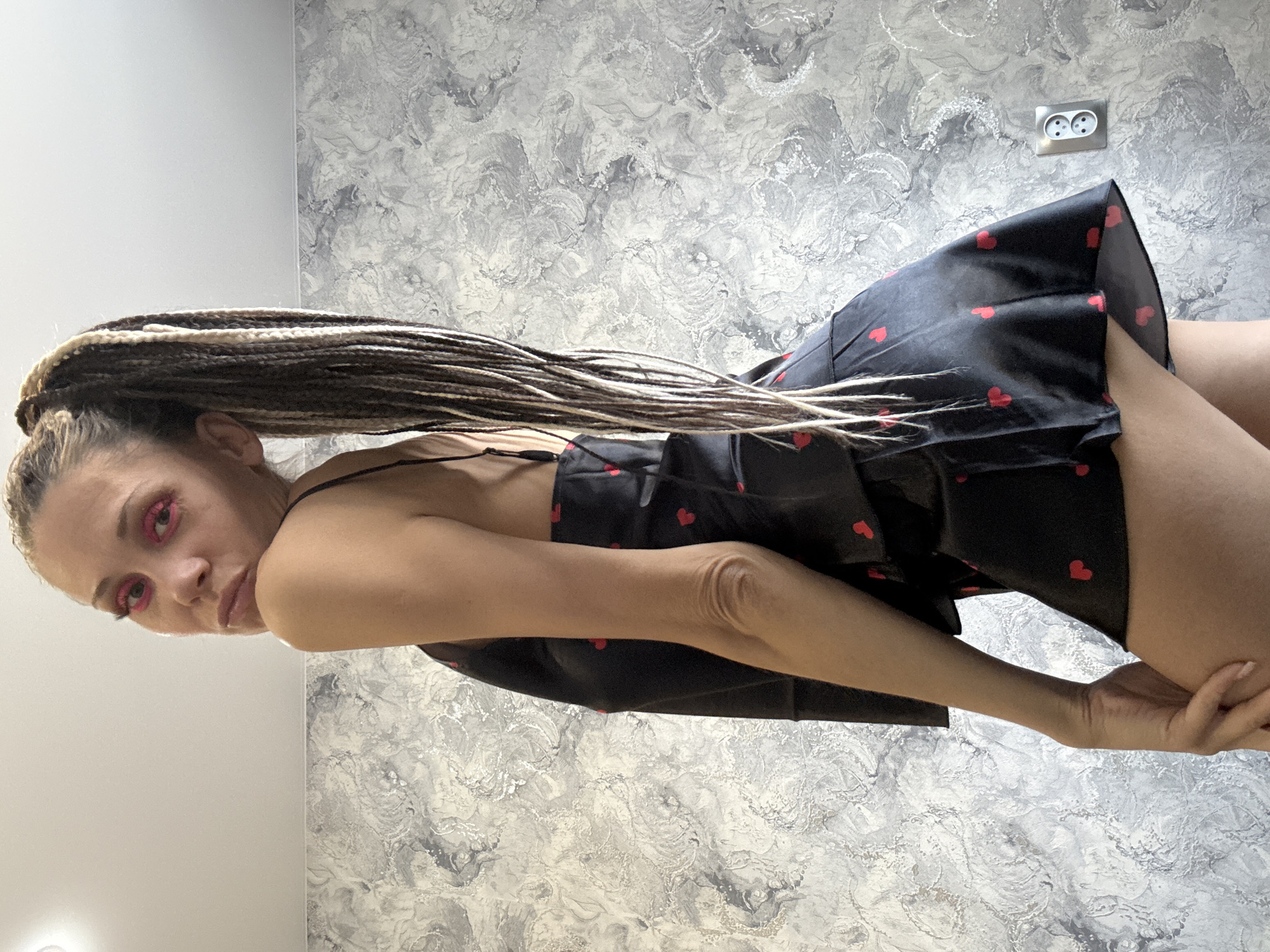 A woman with braided hair is posing indoors. She is wearing a black dress with red heart patterns and pink eye makeup. The background features a wall with a grey marble-like pattern and an electrical outlet. Based on the location provided, the image was likely taken in Rusanovo, Russia.