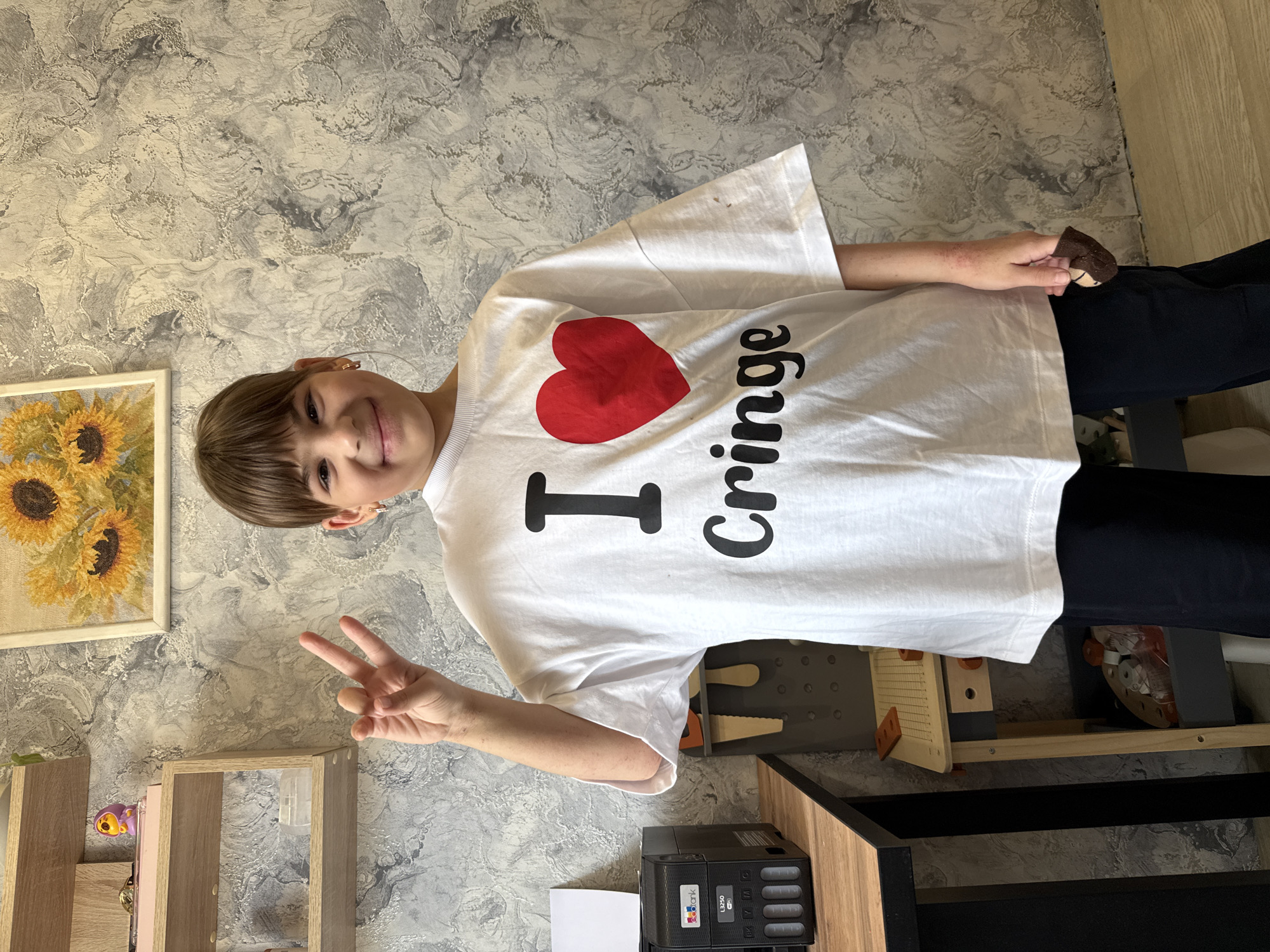 A young girl wearing a white t-shirt with the phrase "I (heart) Cringe" is seen indoors. She is holding up a peace sign with her left hand and appears to be holding something in her right hand. A painting of sunflowers is on the wall to her left. The location is Rusanovo, Russia. Additional details include a wooden shelf, a black printer, and a toy or decorative item.