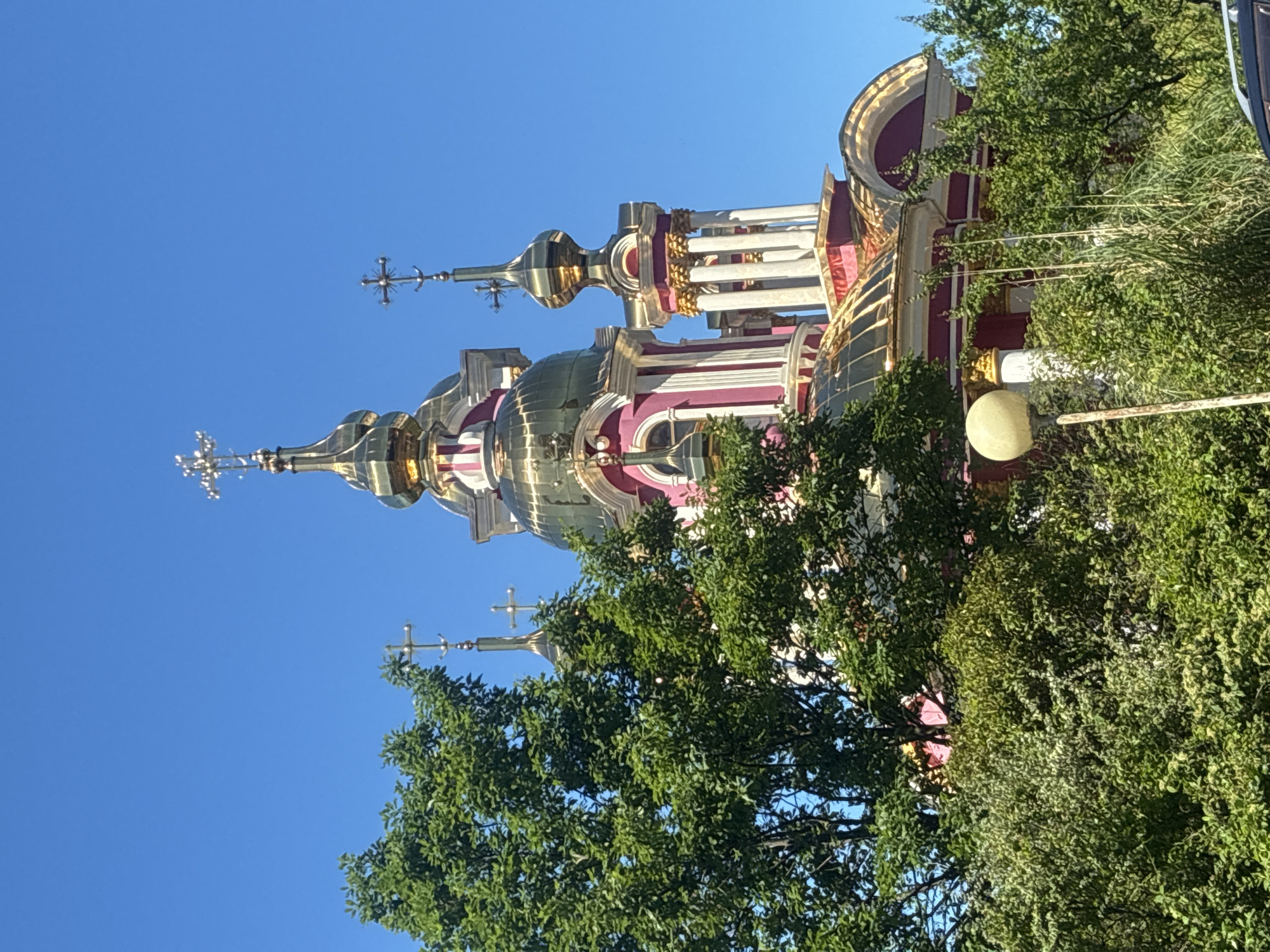 The image displays a vibrant, ornate church building under a clear blue sky, partially framed by lush green trees. The church features multiple gold-colored, onion-shaped domes, each topped with a decorative cross. The main structure of the building is predominantly a rich red or maroon color, accented with white columns and intricate gold detailing along its cornices and decorative elements. Several domes and spires rise towards the sky, showcasing a distinct architectural style, likely Eastern Orthodox.
Dense green foliage from various trees occupies the foreground and middle ground, partially obscuring the lower and right sections of the church, creating a natural frame. The bright sunlight reflects off the golden domes, highlighting their intricate designs. A street light with a spherical lamp and some wires are faintly visible through the leaves on the right side of the image. The clear, bright blue sky suggests a sunny day. No people or specific activities are visible, presenting a serene architectural scene. Given the context, this church is located in Sochi, Russia. No visible text is present in the image.