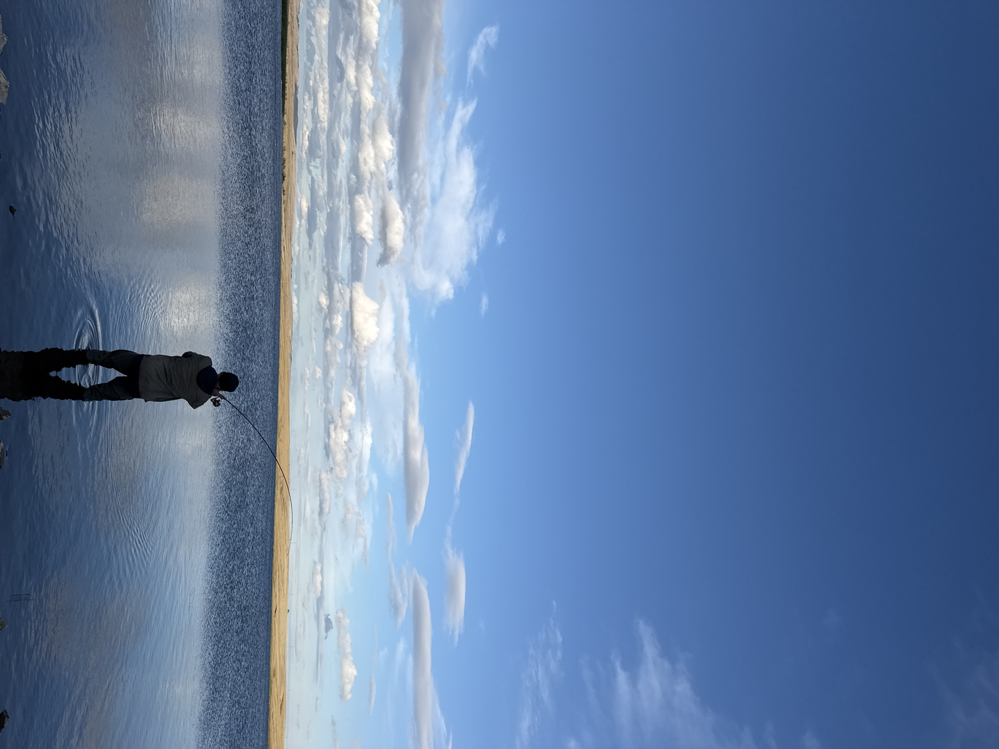 A person stands in a body of water, facing away from the camera, fishing. The person is wearing a jacket and waders. The water reflects the sky, which is blue with scattered white clouds. A flat, dry landscape is visible in the distance. The overall scene depicts a calm and solitary fishing experience in an outdoor environment. The provided metadata indicates the location as Matrayevo, Russia.