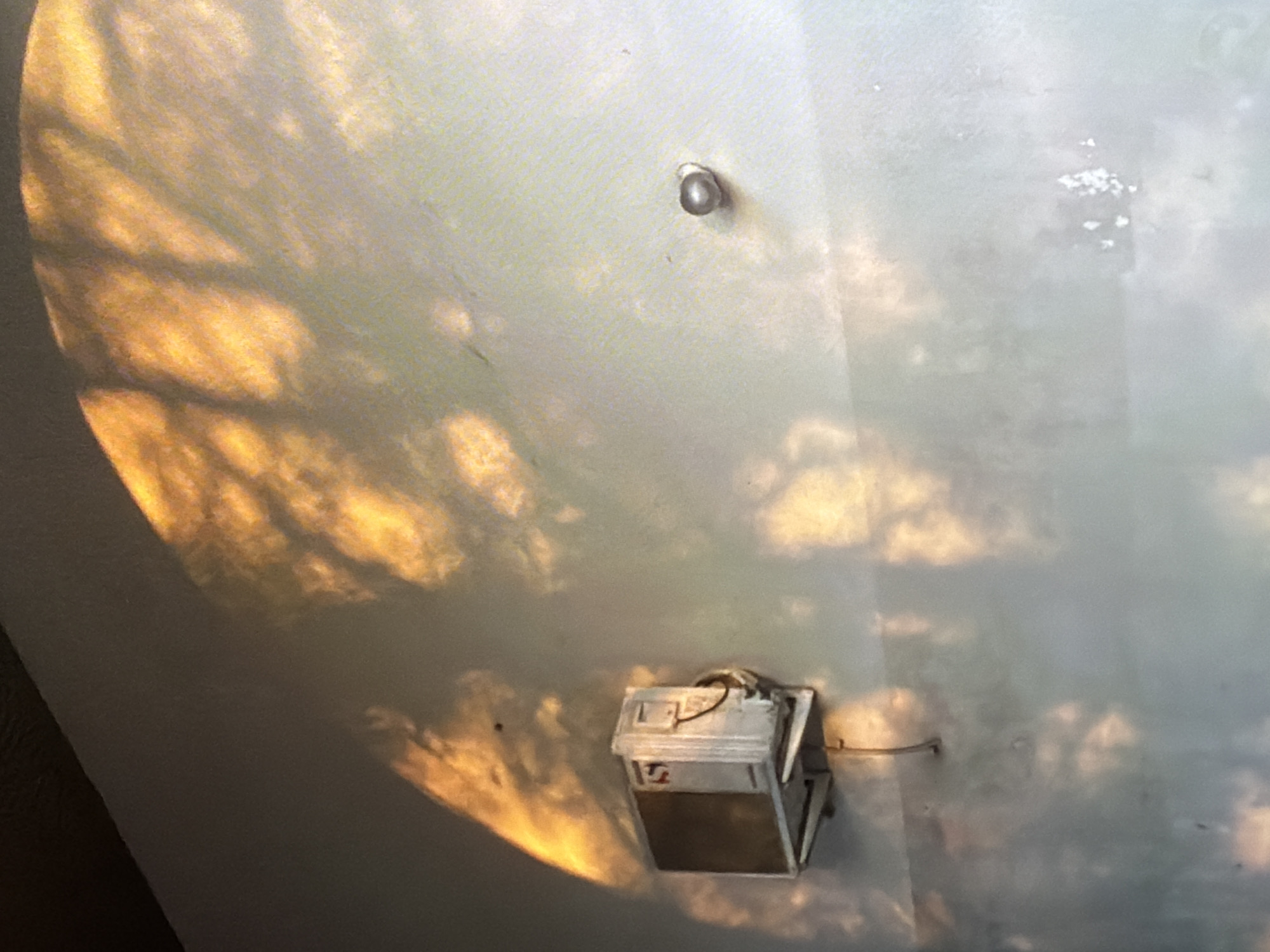 The image depicts an outdoor wall with an air conditioning unit mounted to it. The wall is textured and painted a light color, possibly white or a light gray. Sunlight casts dappled shadows of tree branches onto the wall, creating an abstract pattern of light and dark. The air conditioning unit is a rectangular box with a metallic grille on the front. It appears to be an older model, possibly a window unit installed in a wall sleeve. There is a conduit or wire attached to the unit and running down the wall. To the right of the air conditioning unit, there is a small, round, metallic object attached to the wall, possibly a screw or a small light fixture. The overall scene suggests an urban or suburban setting, likely in a residential or commercial area. There are no people visible in the image.