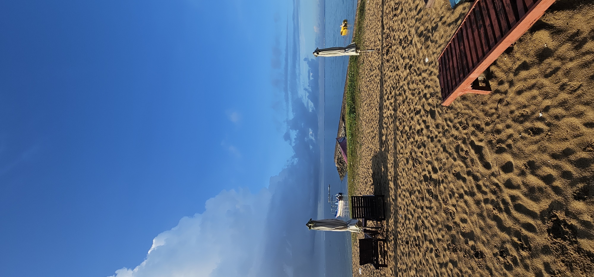 The image depicts a beach in Denpasar, Indonesia. A clear blue sky with cumulus clouds occupies the left side of the frame, contrasting with the brown sand covering the right. The ocean meets the sand along a line where vegetation grows. There are several beach umbrellas and chairs scattered along the sand. A red wooden beach lounge chair is visible in the upper right. A yellow floatation device is in the ocean. A boat and dock are further down the shoreline.