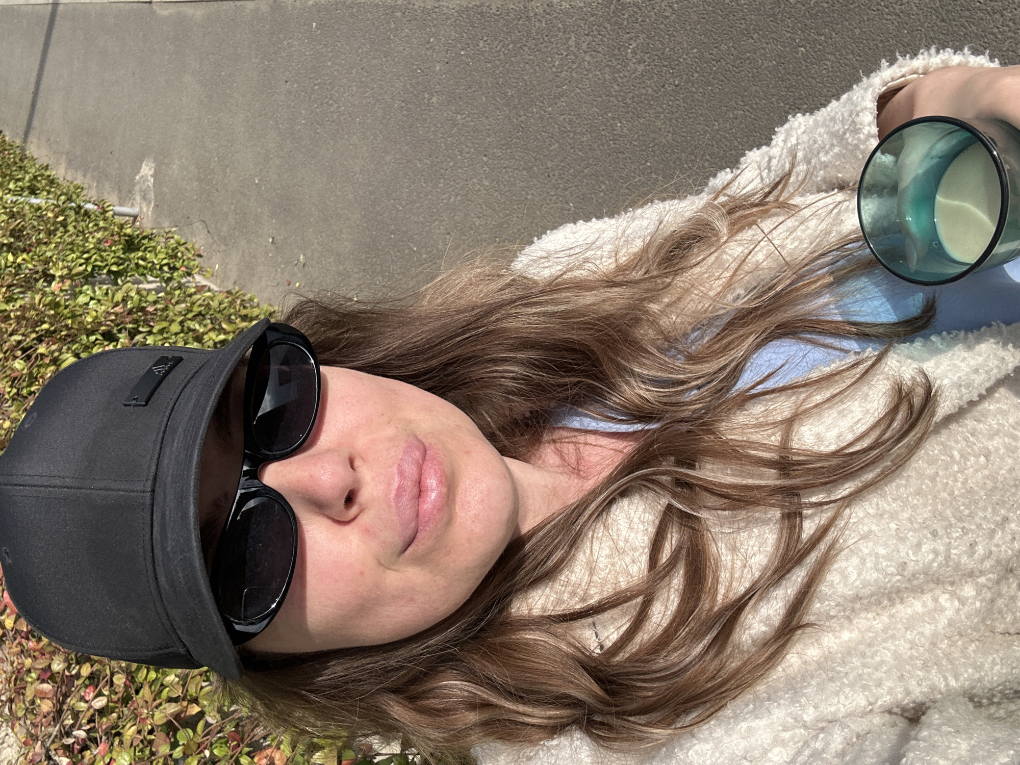 A woman lies on her back in the sunshine. She wears a black baseball cap, large sunglasses, and a white, textured jacket over a light blue top. Her hair is long and brown with light highlights. She holds a teal-colored glass filled with a white liquid. She appears to be resting on an urban surface next to greenery, potentially a sidewalk or a similar public space. The location is not discernible as Tbilisi, Georgia.