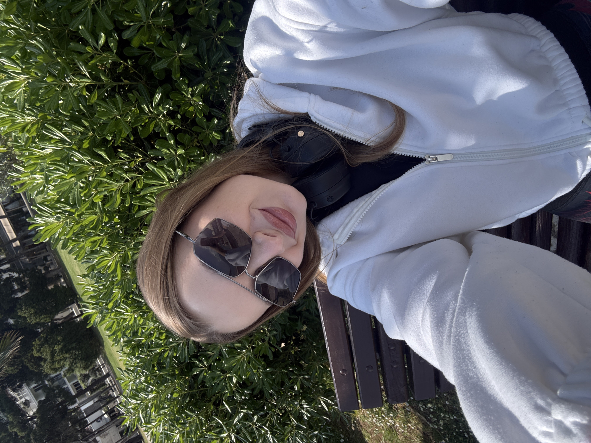 A young person, visible from the chest up, is captured in a brightly lit outdoor selfie. The individual has light brown hair and is wearing large, dark, geometric-shaped sunglasses with a thin metal frame. Their lips are slightly parted, showing a relaxed expression. Around their neck rest black over-ear headphones, partially covered by their hair and a white hooded sweatshirt. The sweatshirt features a silver zipper down the front, and its soft, fleecy texture is evident.
The person appears to be reclining on a dark wooden park bench, parts of which are visible on the right side and below them. The background is dominated by dense, lush green foliage, characteristic of bushes or trees, indicating a natural outdoor setting like a park or garden. In the upper left background, some light-colored buildings and more distant greenery can be seen, suggesting an urban or suburban environment. The ground beneath the bench shows patches of small white flowers or details.
The scene is bathed in strong, natural sunlight, indicating it is daytime with clear weather. The overall impression is one of relaxation and comfort while enjoying the outdoors in Budva, Montenegro. There is no visible text in the image.