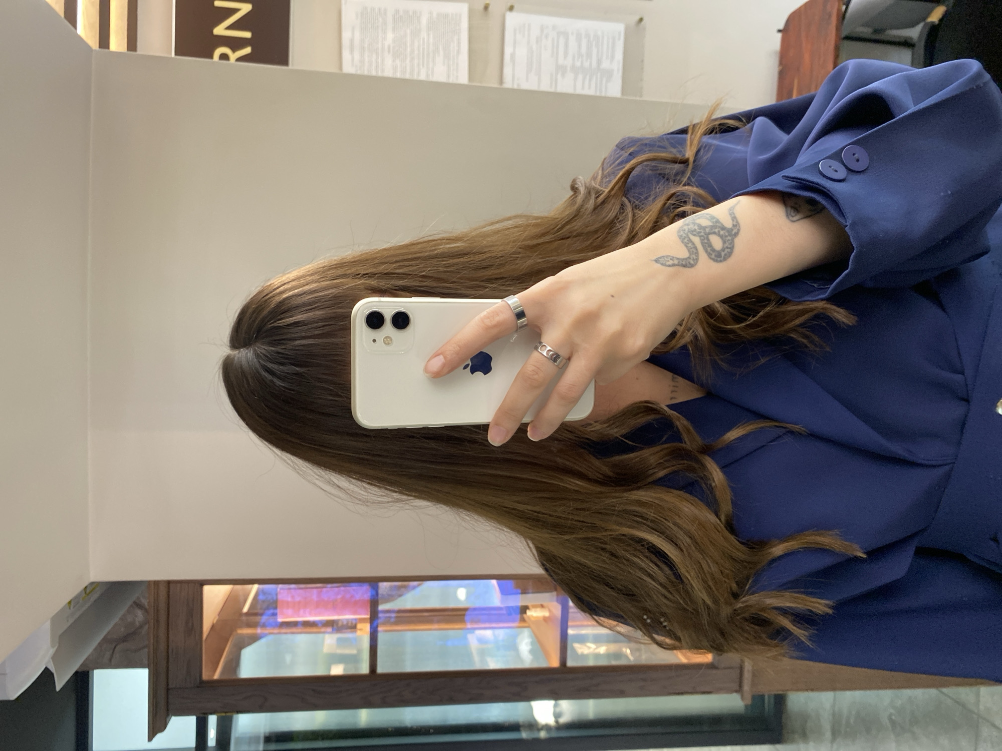 A young woman with long, brown hair takes a mirror selfie holding a white iPhone. She is wearing a dark blue blazer and several silver rings on her left hand. A snake tattoo is visible on her forearm. The background appears to be an indoor space with a wooden cabinet to the left and framed documents on the wall to the right. The image does not provide any information about the city of Votkinsk, Russia.