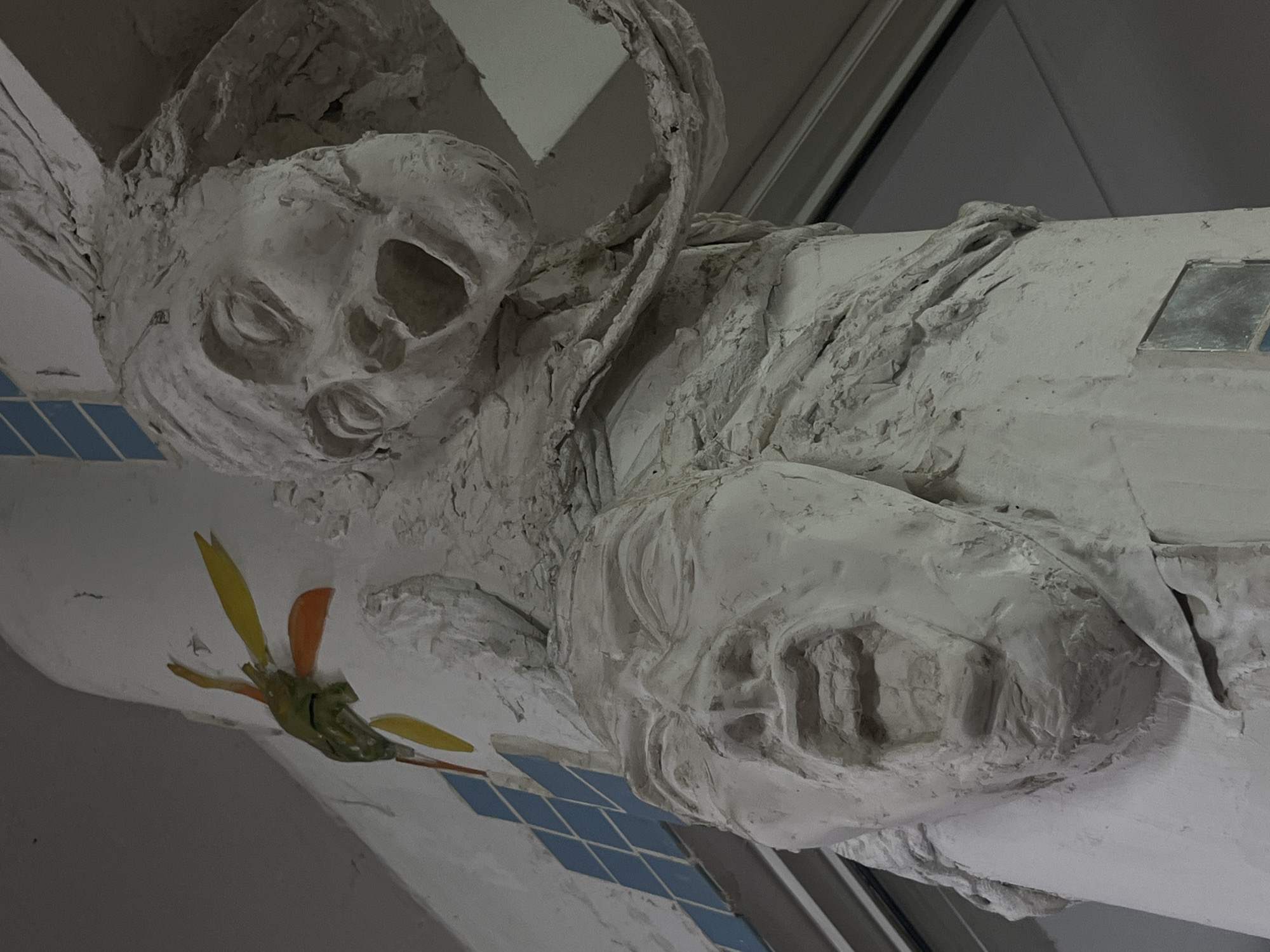 The media file presents a close-up view of a large, highly textured, white sculpture integrated into an architectural element, likely a beam or support structure, within a building in Buenos Aires, Argentina. The sculpture depicts a reclining human-like figure with a grim, skull-like face. Its features include deep, hollow eye sockets, a prominent bony nose, and an open mouth revealing teeth, giving it a weathered, eerie, or solemn appearance. Roughly sculpted, wing-like or drapery-like forms extend behind and above the figure's head.
The sculpture's body, characterized by its rough, uneven surface, appears to merge seamlessly with the supporting structure. This main structure is primarily white with a similar rugged texture. Adorning the structure below the sculpture are light blue square tiles arranged in a grid pattern. Further along the white surface, a small, vibrant decorative motif resembling a stylized hummingbird or butterfly is visible, crafted from yellow, orange, and green pieces. A rectangular glass or mirrored inlay is also present on the structure.
The overall impression is that of a static artistic embellishment or an architectural detail with an ancient or decayed aesthetic. The lighting is soft and diffused, suggesting an indoor or covered outdoor setting without strong shadows. No specific activity or event is taking place. There is no visible text in the image.