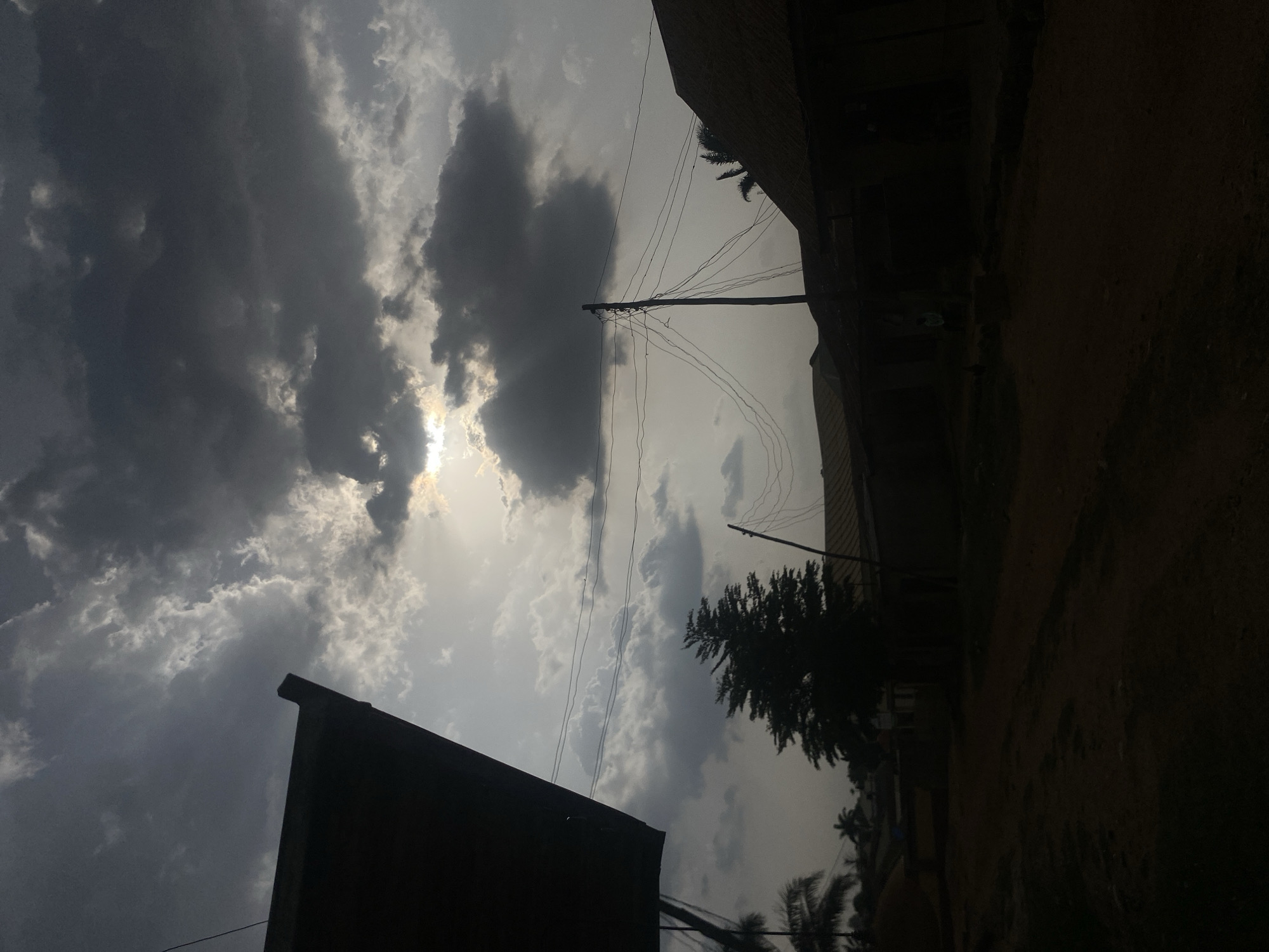 The image shows a view of Narayi, Nigeria, under an overcast sky. Predominantly gray clouds fill the sky, with a patch of sunlight breaking through. Power lines run horizontally across the frame, connecting to wooden utility poles. A tree with dense foliage is visible. Silhouetted buildings are positioned on the right side of the image, obscuring much of the foreground. A dark roof is visible in the lower left corner of the frame. The ground is dimly lit, blending into the darkness on the right side.