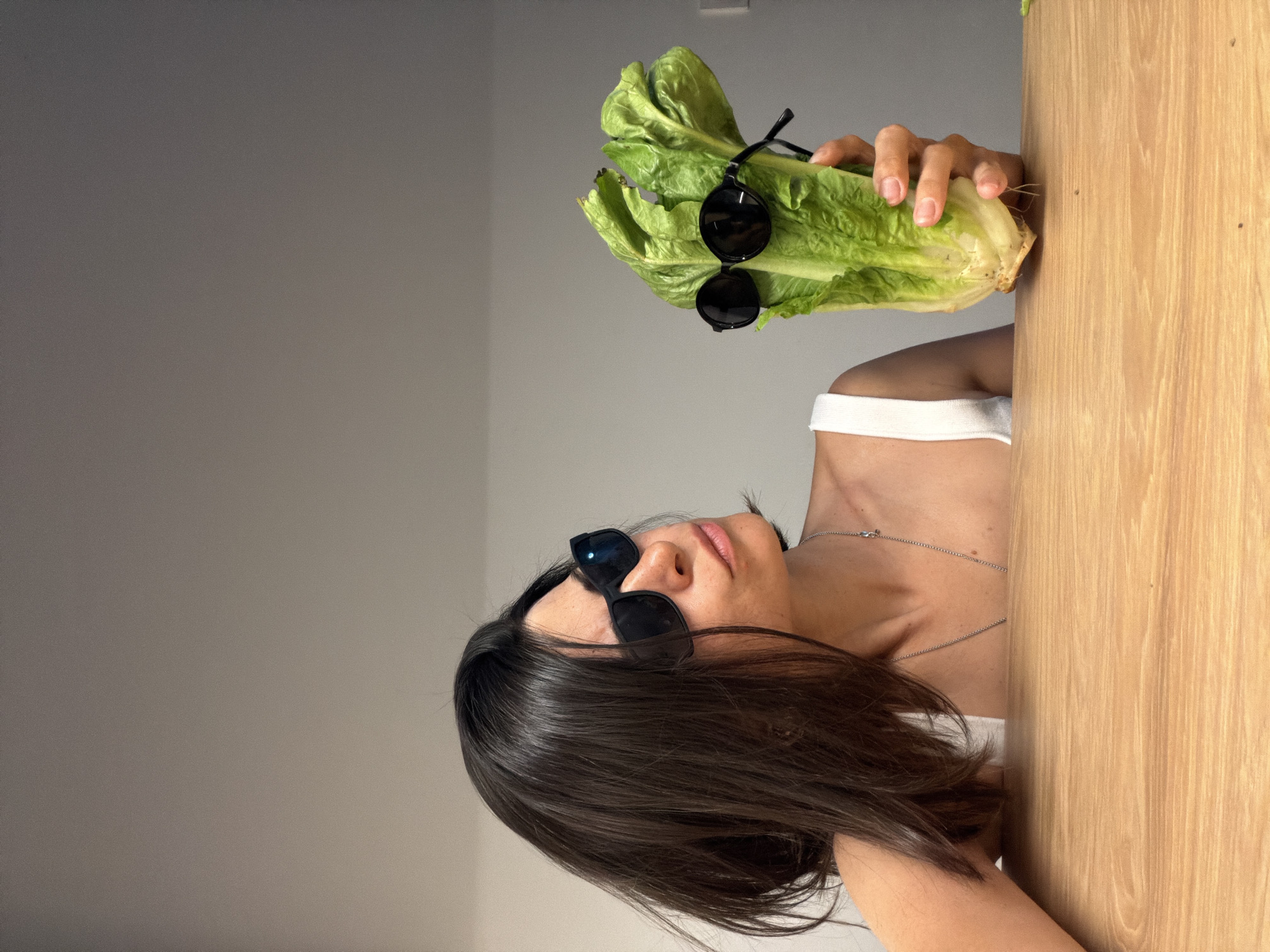 A person wearing sunglasses and a white tank top is holding a head of romaine lettuce with sunglasses on it, next to a light wooden cabinet or wall. The setting appears to be indoors against a light gray wall. The location is not identifiable as being in Budva, Montenegro from the visual data provided.