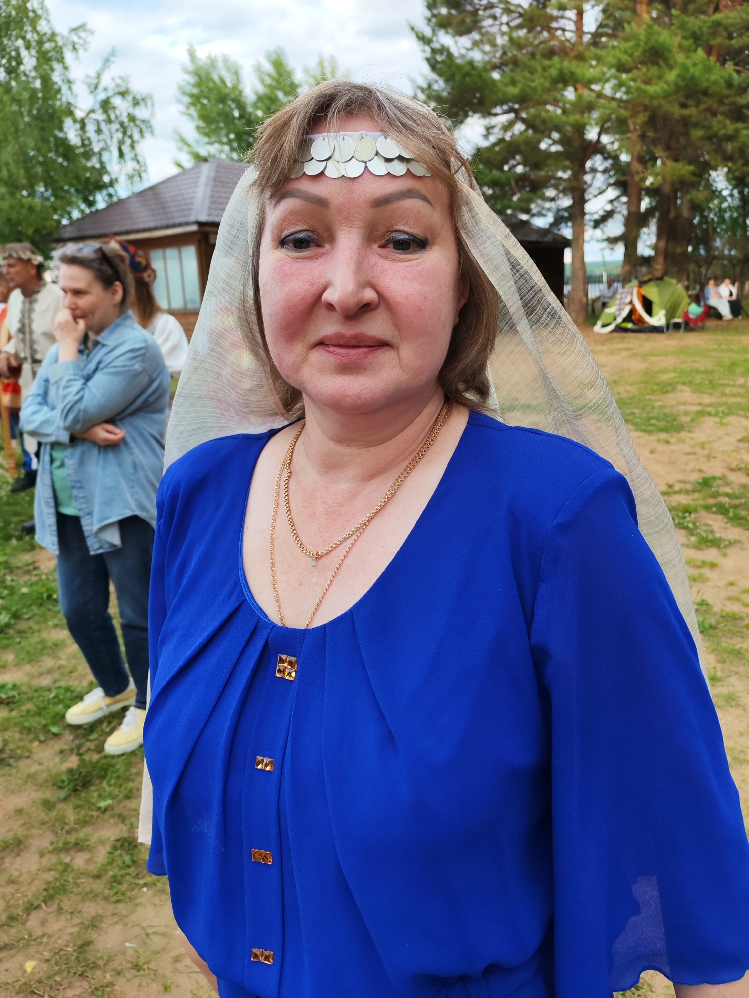 A woman, wearing a blue dress and a headdress adorned with silver coins, stands in the foreground of an outdoor event. She is looking directly at the camera with a neutral expression. Behind her, several other people are visible, some dressed in traditional clothing. In the background, there's a wooden building, trees, and tents, suggesting a festival or gathering. The lighting indicates it's daytime, possibly late afternoon given the soft light. The setting appears to be a grassy area with dirt paths, consistent with an outdoor park or festival grounds in Chaikovsky, Russia. The woman's attire, particularly the headdress and the decorative buttons on her dress, suggest a cultural or historical theme to the event.