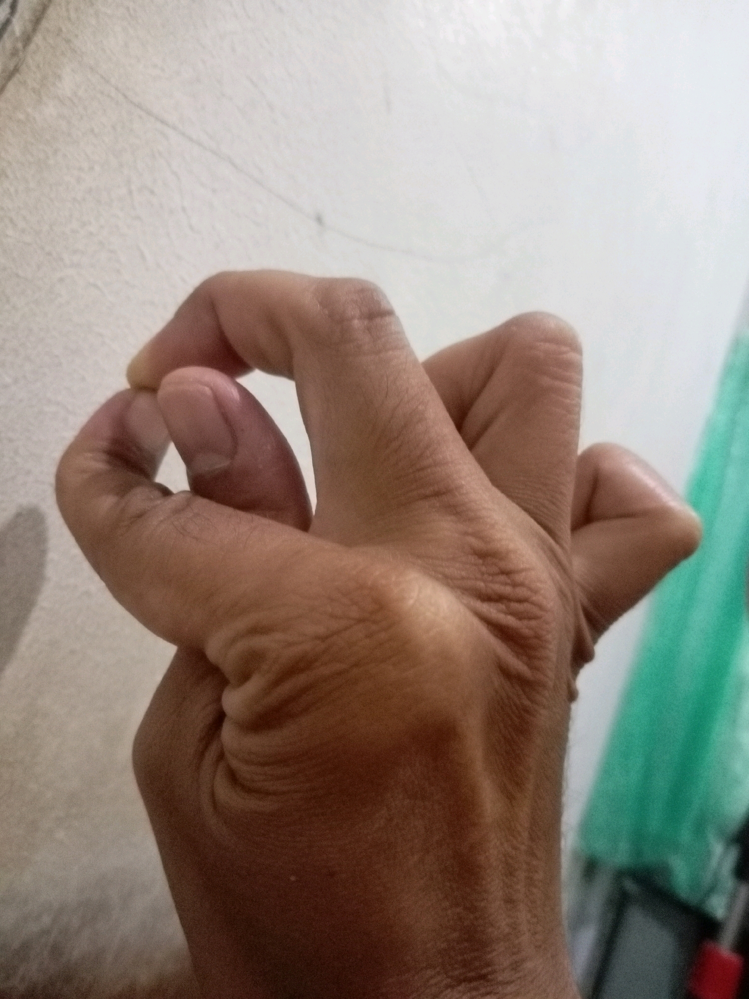 A close-up, medium-angle shot shows a person's right hand, with fingers bent and intertwined in a way that suggests a grip or a gesture. The hand is positioned against a textured, off-white wall, with a blurry green object and a black object with a red accent visible in the background on the right side. The lighting appears to be from the left, casting a shadow of the hand on the wall. The image was taken in Batam, Indonesia.