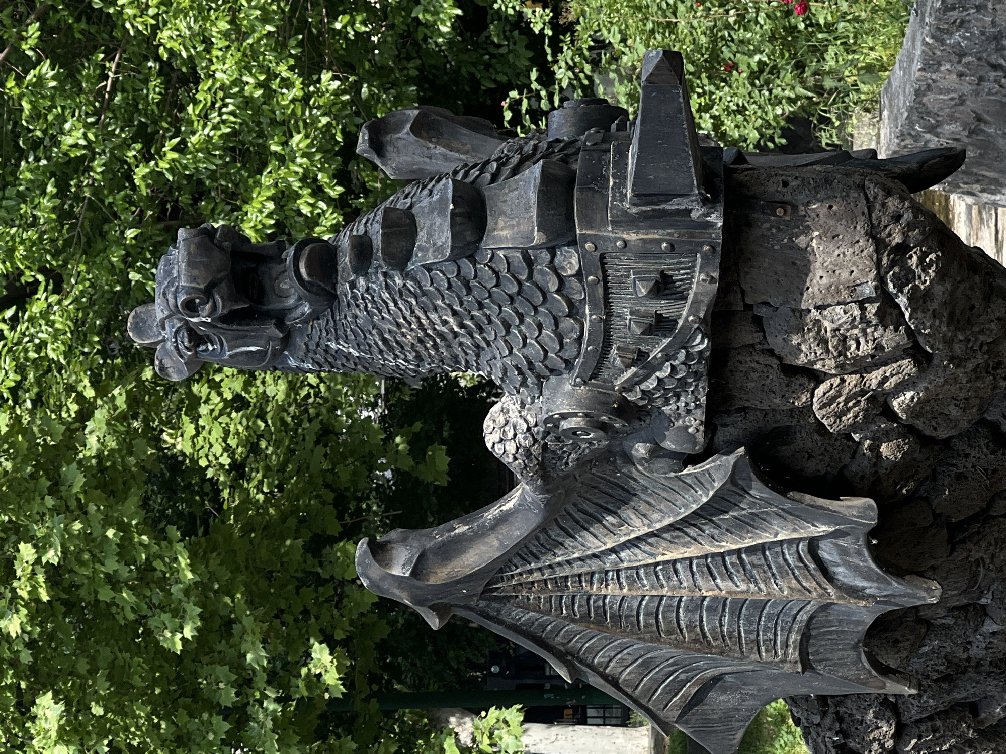 A vertical, medium shot depicts a metal sculpture of a dragon, possibly made of dark bronze or a similar material. The dragon's head is on the left side of the image with its wing stretched downward. The dragon has a scaly texture across its body and intricate details in its wings. Part of the sculpture is attached to a darker rock formation. The background is filled with green foliage, suggesting the sculpture is located outdoors, possibly in a park or garden. The location is in the city of Unknown, Unknown.