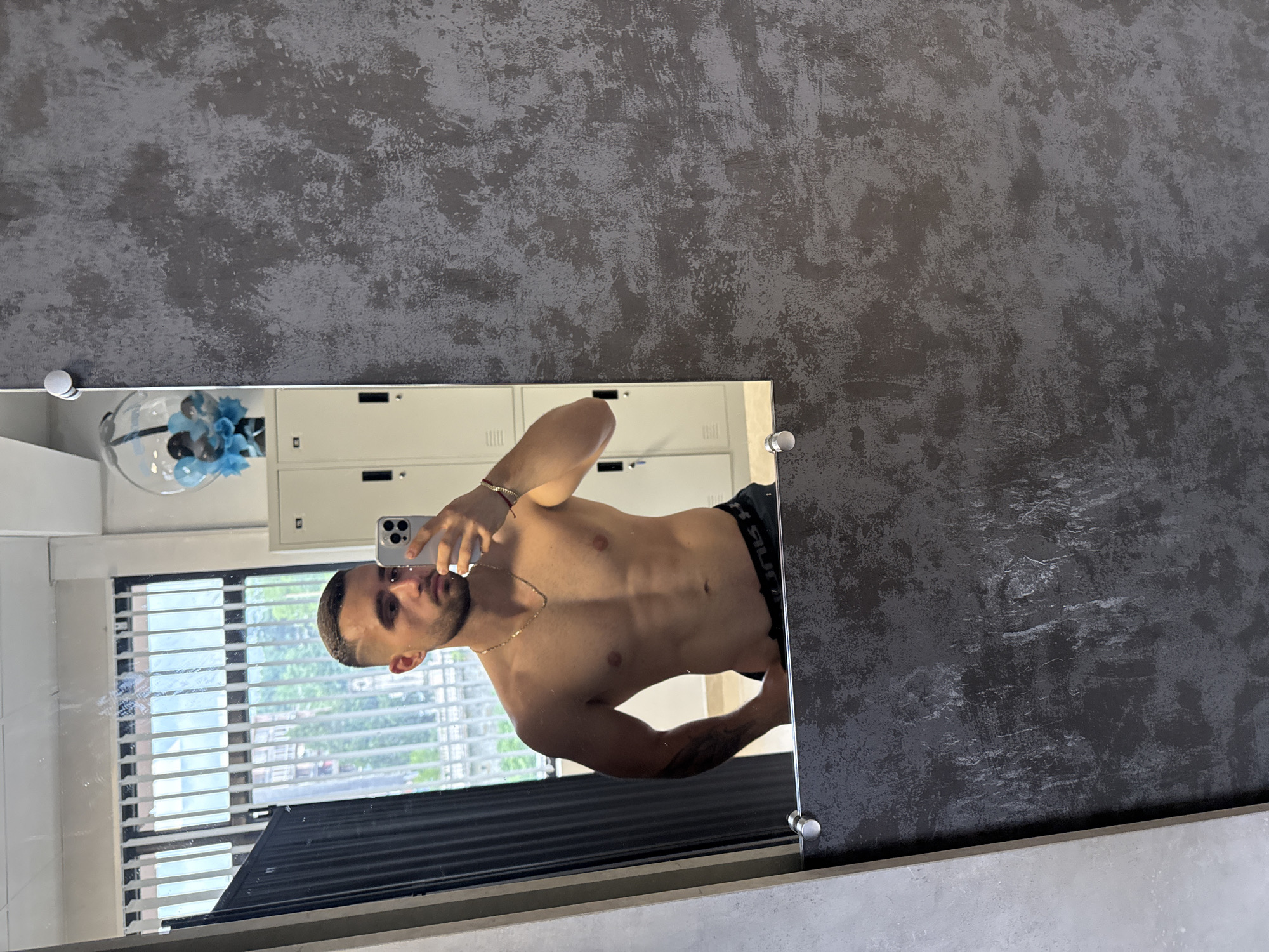 A shirtless man with short, dark hair takes a mirror selfie with a smartphone. He stands in what appears to be a locker room, based on the row of lockers visible behind him in the mirror. Outside the window, through the blinds, there is a view of buildings and greenery, typical of a city landscape. The mirror is attached to a wall with a textured, dark gray finish. The man wears a gold chain and a bracelet. The image does not provide any clues about the location in Gabrovo, Bulgaria.