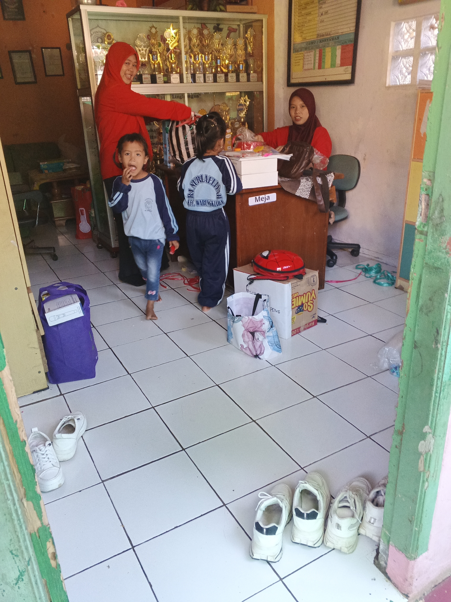 In Warungkiara, Indonesia, a scene unfolds within what appears to be a school or community center. Two women, both wearing hijabs, are present. One woman, in a red hijab and red shirt, stands near a display cabinet filled with trophies, interacting with a young boy in a blue and white uniform who is looking at the camera with his hand near his mouth. The other woman, in a dark hijab and red shirt, sits behind a wooden desk labeled "Meja" (Table), appearing to be working or assisting.
A young girl, also in a blue and white uniform, stands between the two women, facing away from the camera. In the foreground, a purple tote bag rests on the tiled floor, and a collection of white sneakers is arranged near a doorway framed by green and yellow elements. Boxes and bags are scattered around the desk area, suggesting an ongoing activity or transaction. The environment is brightly lit, with tiled floors and walls visible throughout.
