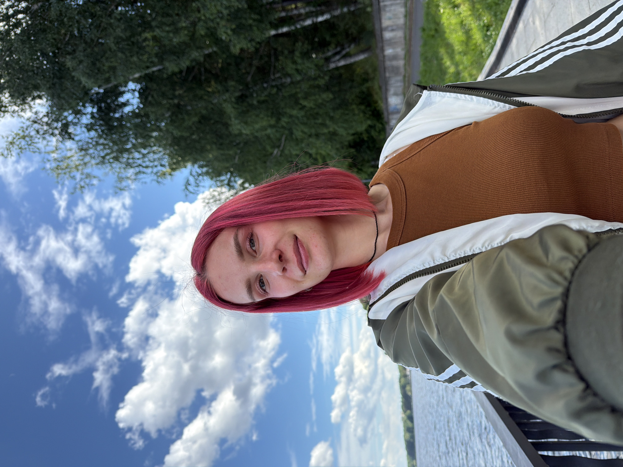 A young woman with short, bright pink hair and a tan ribbed tank top is posing for a selfie. She is wearing an olive green bomber jacket with white stripes on the sleeves and a white lining. The background shows a body of water, possibly the Kama River, and a line of trees on a sunny day with clouds in the blue sky. The location is along the embankment of the city of Chaikovsky, Russia, where a paved walkway is visible.
