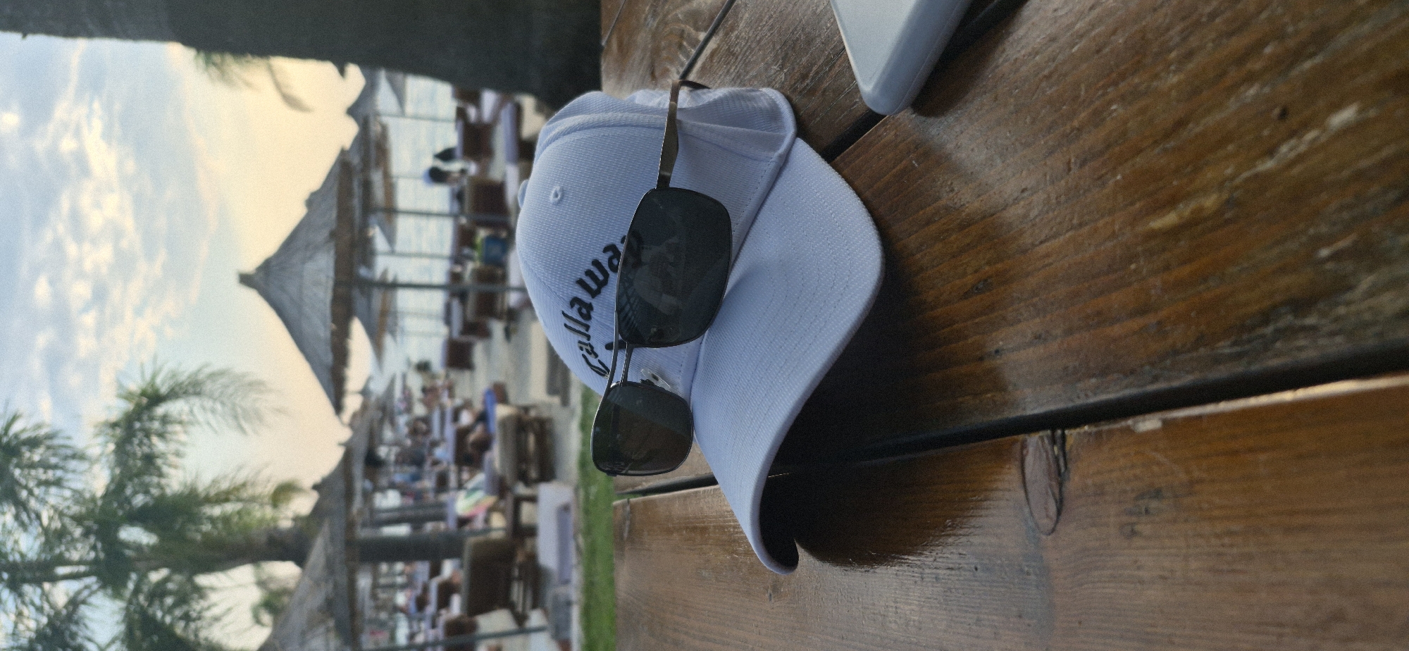 The image depicts a close-up view of a white Callaway golf cap with dark sunglasses resting on top, placed on a wet wooden table. In the background, a beach scene unfolds with numerous thatched umbrellas and lounge chairs. Palm trees are visible, suggesting a tropical or coastal setting. The sky is partly cloudy with soft hues of orange and blue, indicating either sunrise or sunset. The beach appears to be populated with several people relaxing or strolling. The scene is likely set in Saranda, Albania, a city known for its beaches and coastal scenery. The lighting suggests it's an outdoor setting during a warm part of the day.