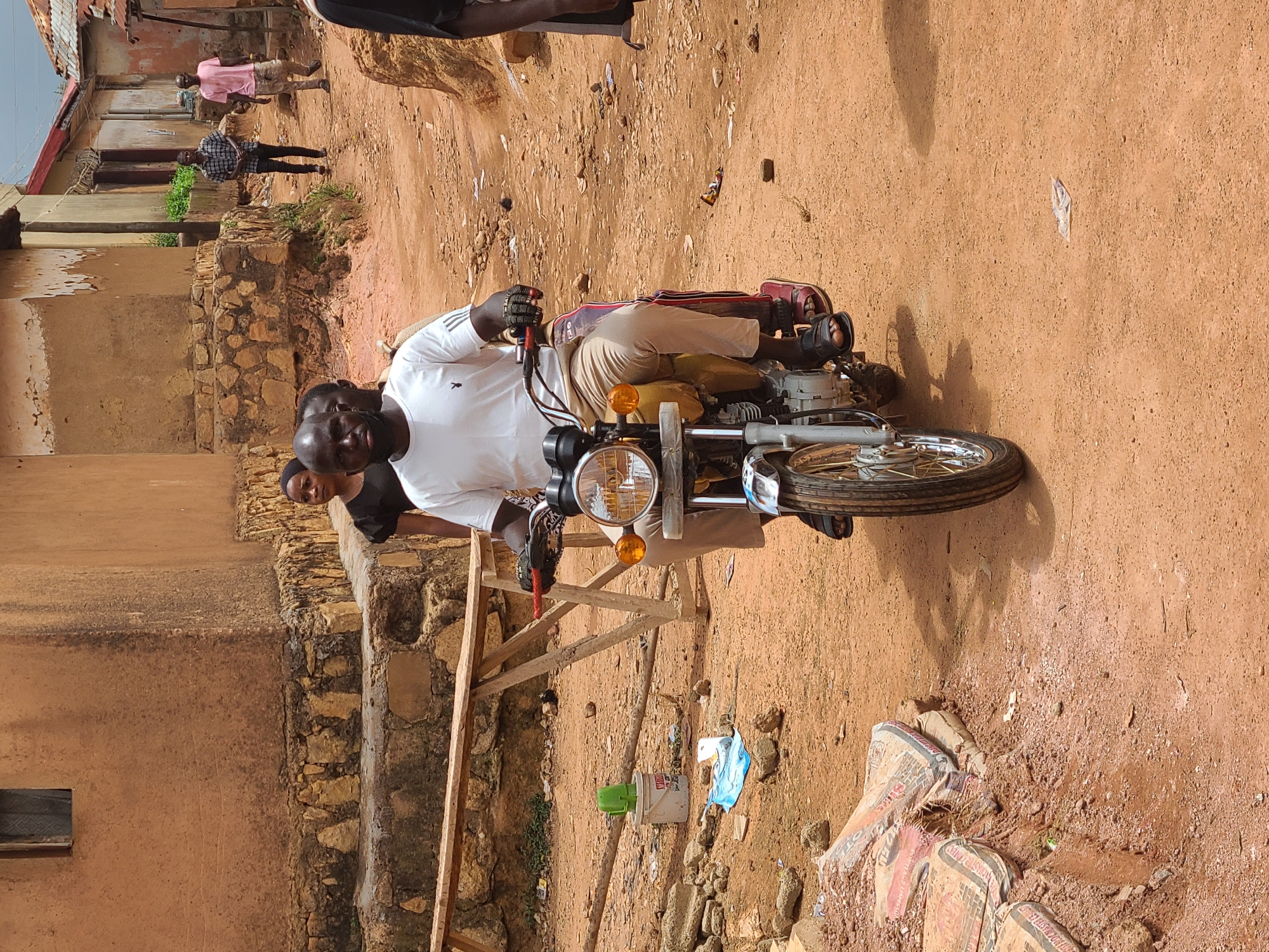 A medium, slightly rotated, shot depicts a man in a white t-shirt straddling a motorcycle in an outdoor setting. The ground appears to be reddish-brown dirt with scattered debris and rocks. Behind the man, there is a partially visible stone or brick structure. A wooden structure stands nearby. In the background, there are blurred figures of people walking in the distance. A white bucket and blue plastic bag lie on the ground. Cement bags are in the lower right.