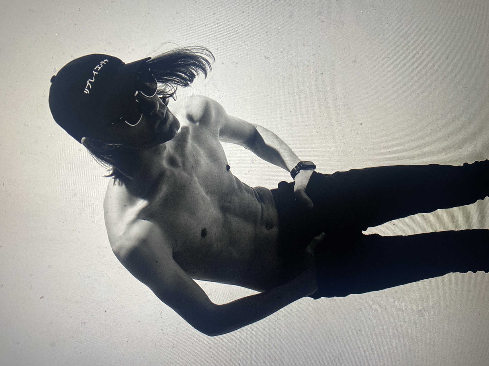 A black and white photograph depicts a shirtless individual with dark hair, a cap with text in a foreign language, dark pants, and sunglasses. They are wearing a watch and an earring. The background is a solid white tone. The photo's angle is oblique, suggesting the subject is positioned above the viewer. There is no evidence that this image was taken in the location of Шебунино, Russia.