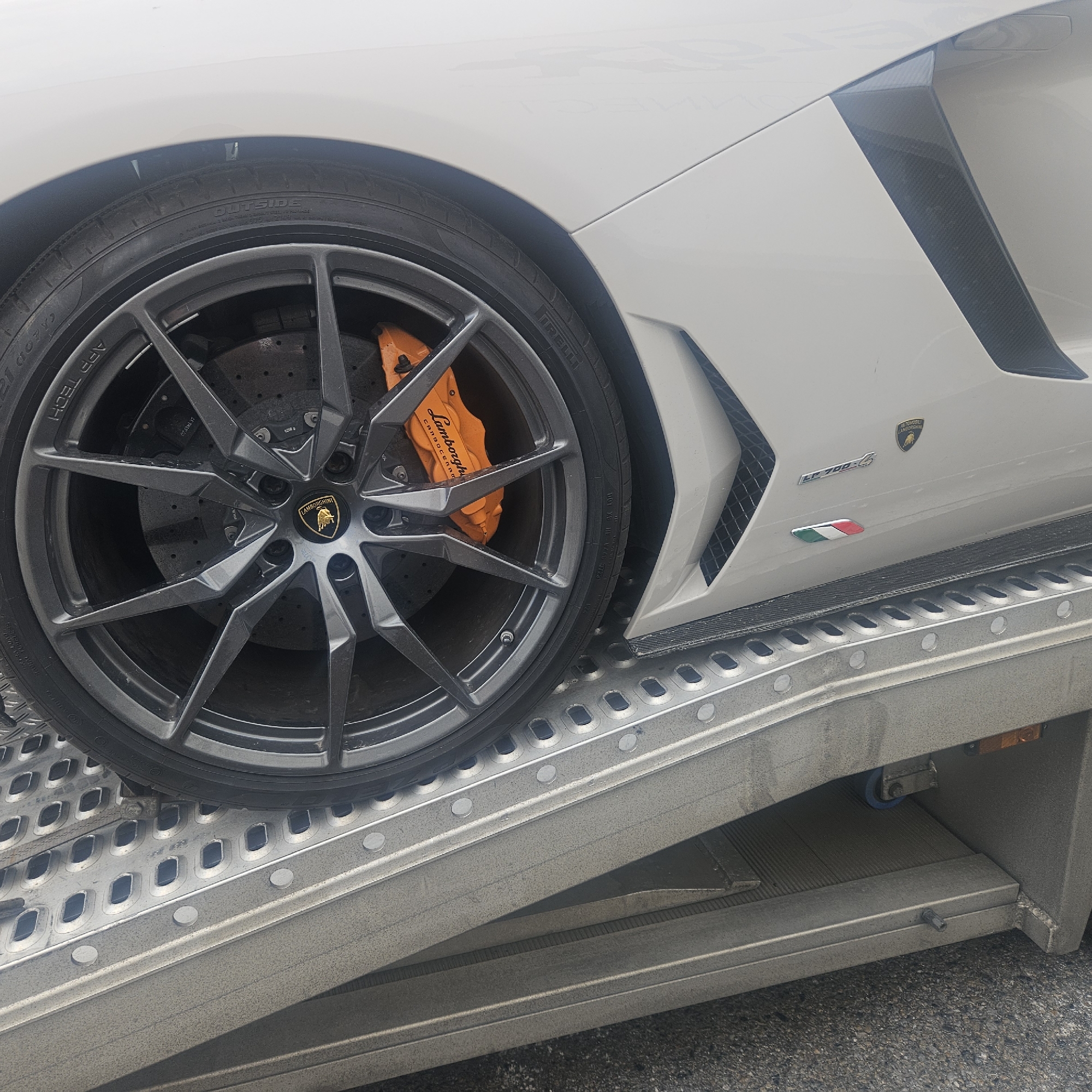 The image shows the front passenger-side wheel and part of the body of a white Lamborghini Aventador, with the Italian flag decal visible on the side. The car is positioned on a silver ramp, which is part of a trailer or truck, likely for transport. The ramp's surface is textured and metallic. The background is blurred, suggesting an outdoor setting. There are no people visible in the image. The scene appears to be in La Jonquera, Spain, given the context of the car being on a transport ramp.