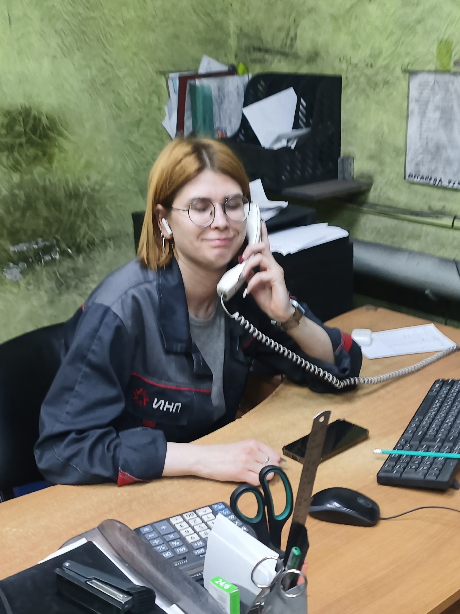 A woman with auburn hair is sitting at a desk, talking on a corded telephone. She wears wire-rimmed glasses and a gray uniform jacket with a red stripe and the Russian letters "ИНП" on the left breast. An earpiece is visible in her left ear. The desk is cluttered with office supplies, including a calculator, a stapler, scissors, a ruler, a mouse, a keyboard, a pencil, a smartphone, and paper. The background is a green wall with various items hanging on it. Based on the text on the items, this is in Noviy, Russia.