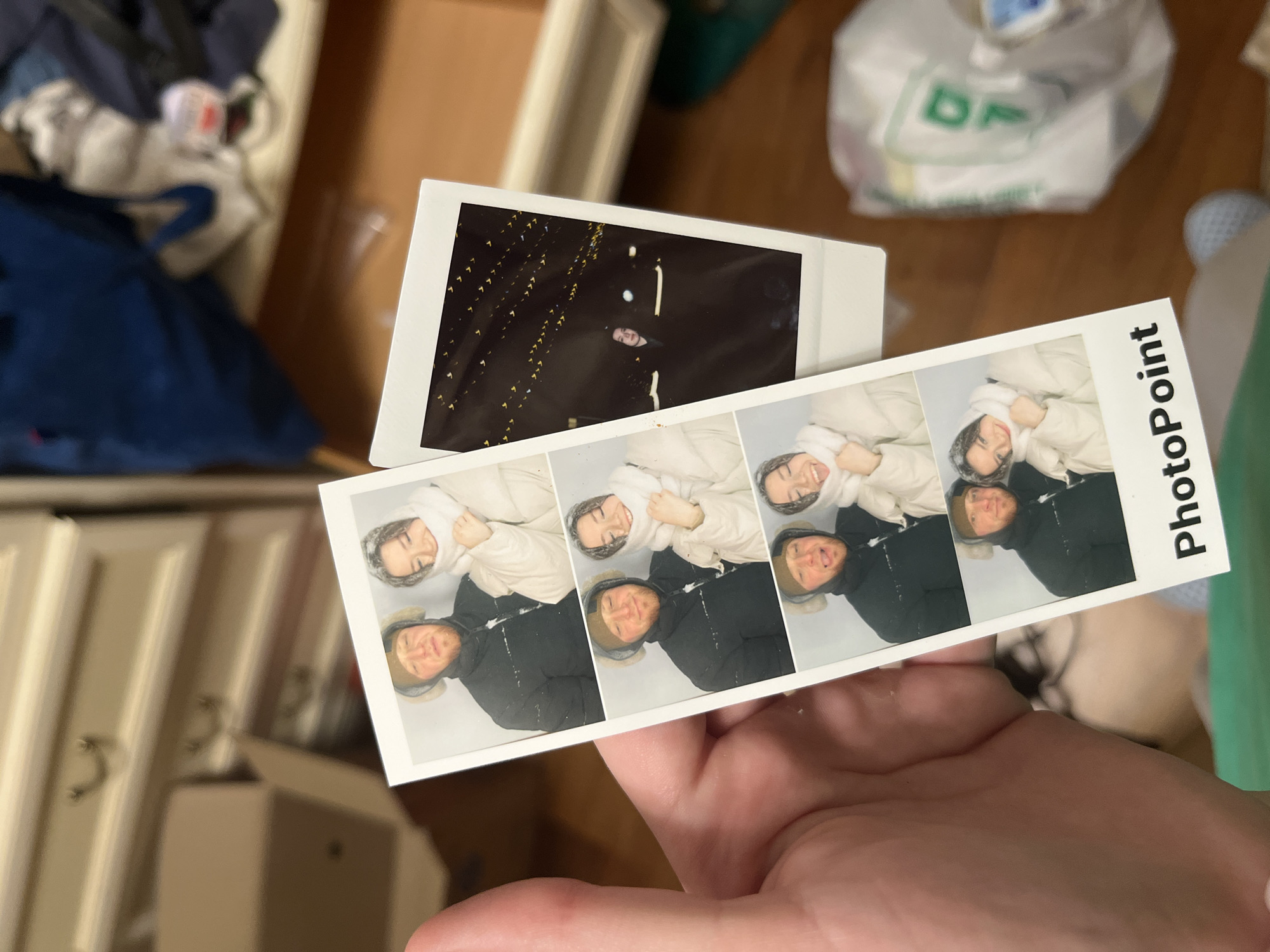 The image shows a hand holding two photographs. One is a photo booth strip from PhotoPoint, showing four images of two people wearing winter clothing, likely taken outdoors in snowy conditions. The other is a Polaroid-style photo of a person standing in front of illuminated lights, potentially taken outdoors at night. The background includes furniture, a blue garment, and a white plastic bag with green lettering. There is no information to determine the location of the photographs to be in Moscow, Russia.