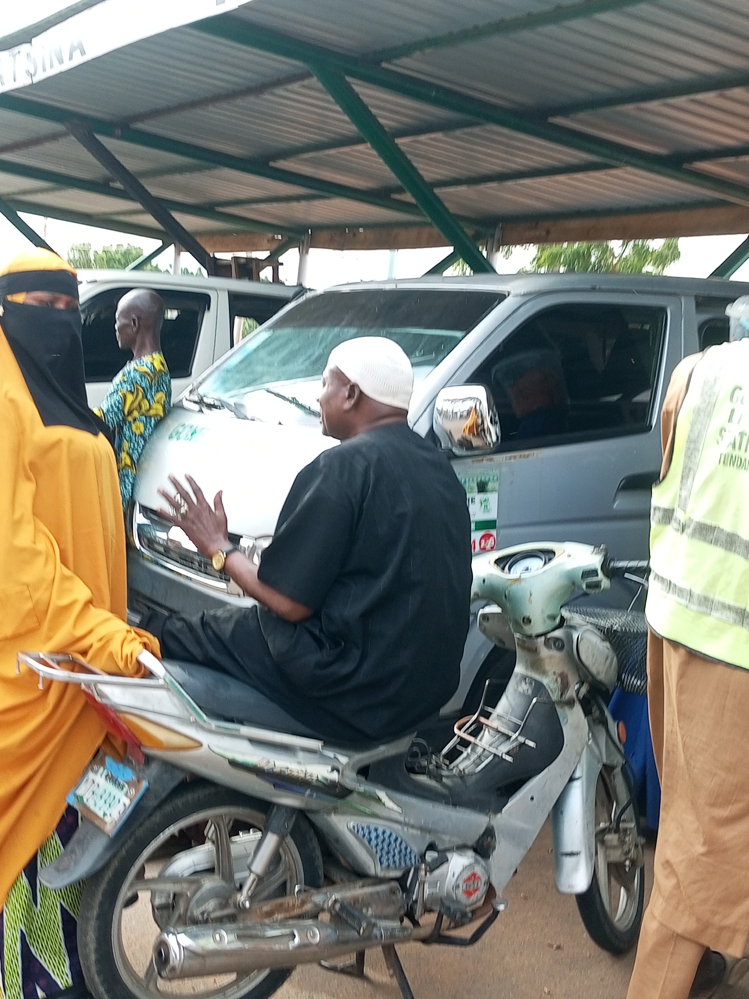 In Gombe, Nigeria, under a corrugated metal roof supported by green metal beams, a group of individuals are gathered near a silver van. A woman in a bright yellow hijab and black face veil stands on the left, partially obscuring a white van behind her. To her right, a man in a colorful African print shirt stands facing away from the camera. In the foreground, a man wearing a white kufi and a black thobe sits on a silver and light green motorcycle, gesturing with his right hand. His left hand is on the handlebars. The motorcycle has a white license plate. To the right of the man on the motorcycle, another person, partially visible, wears a bright yellow safety vest over a brown garment. The ground appears to be dirt or gravel. The overall scene suggests a public gathering or transportation hub.