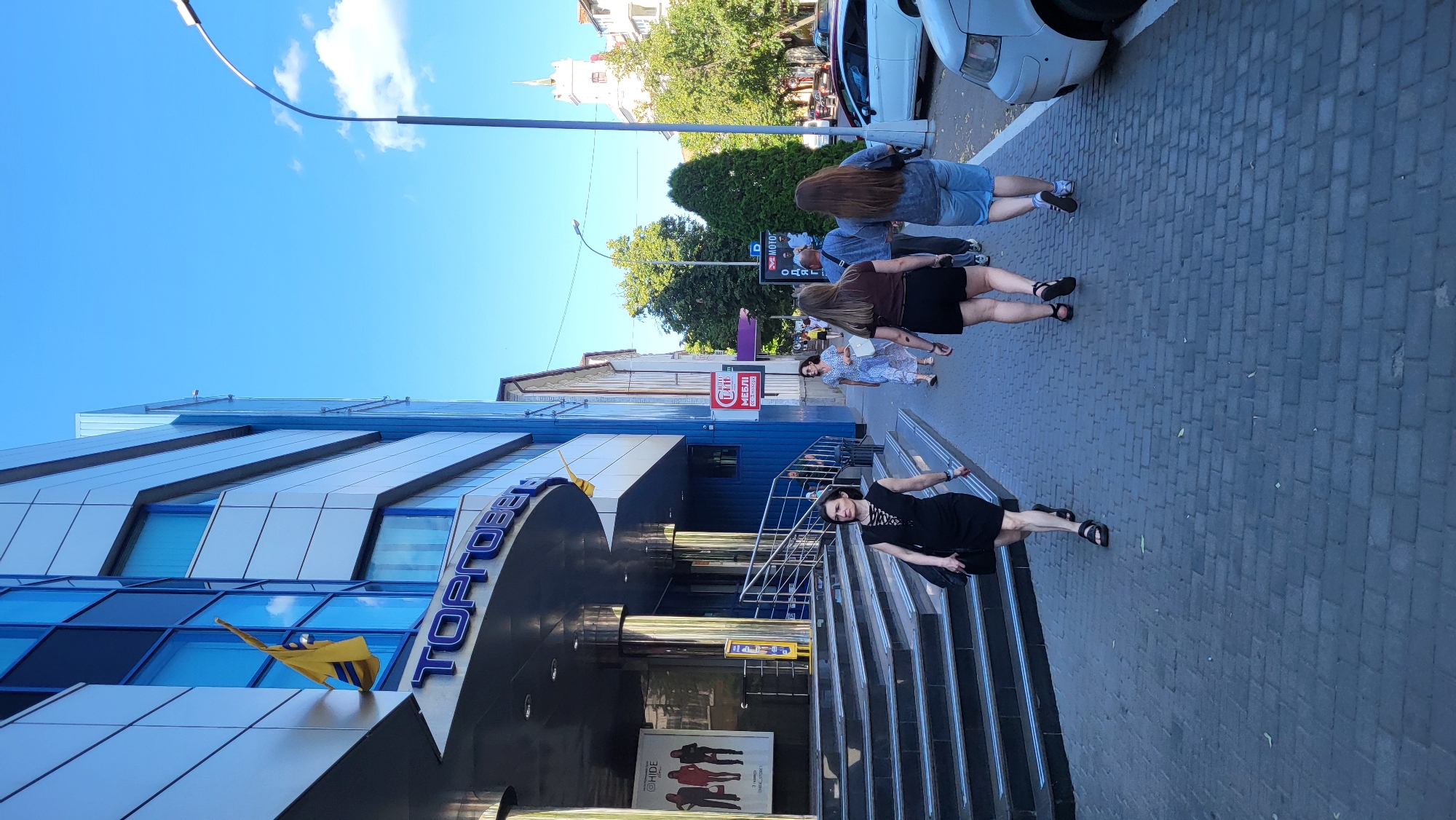 In a bright, sunny scene, a group of people walk along a paved sidewalk in front of a modern, blue and white building. The building has a large, blue sign with white lettering that reads "ТОРГОВЕ". A Ukrainian flag, yellow and blue, is displayed on a pole attached to the building. A woman in a black dress walks down a set of wide stairs leading away from the building. Other pedestrians, including two women in the foreground walking away from the camera, are visible on the sidewalk. To the right of the building, a street with parked cars and trees lines the area, with a tall tower visible in the distance. The sky is clear blue with a few white clouds.
