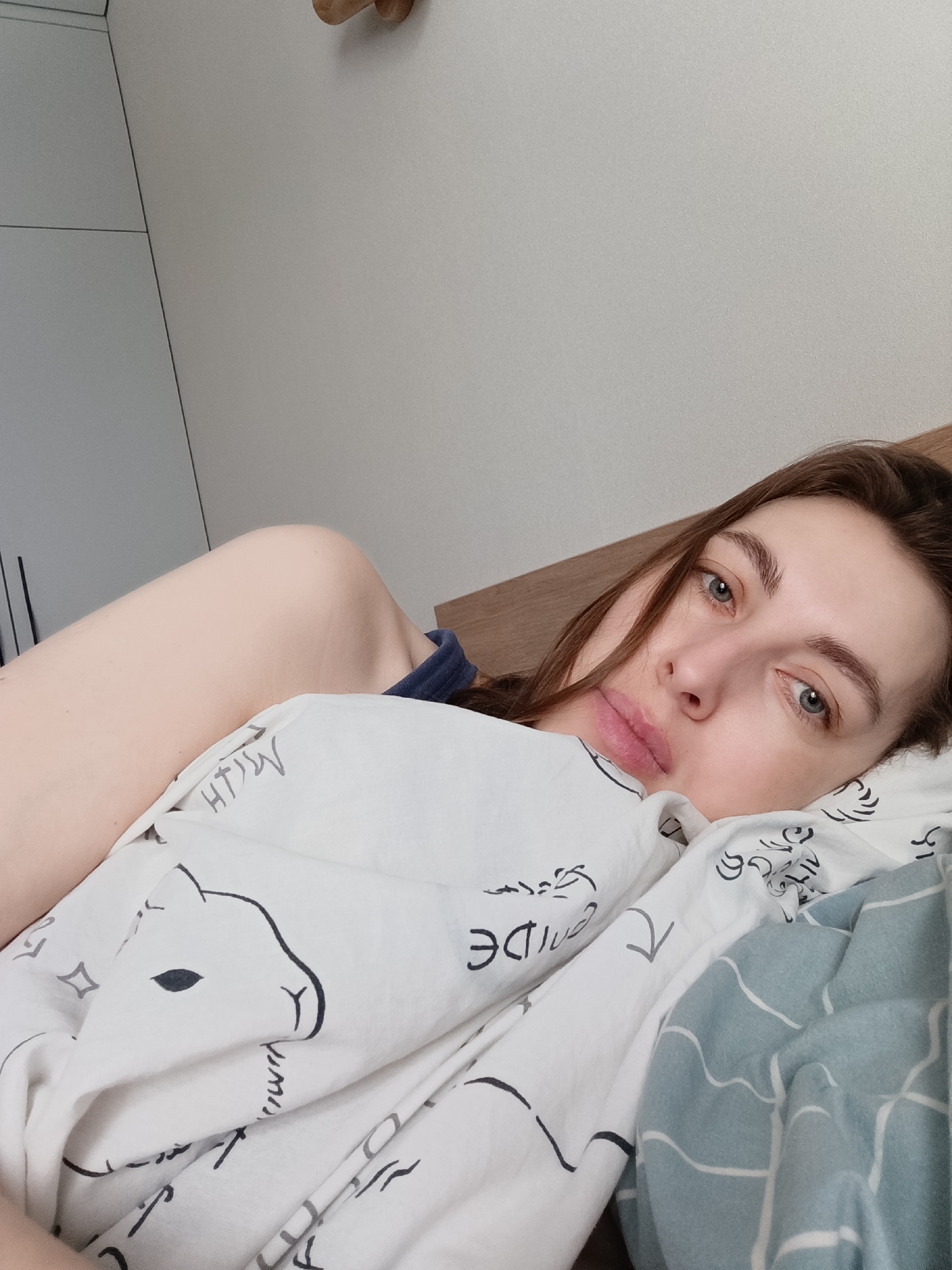 An adult female is depicted lying in a bed indoors, located in the city of Chaikovsky, Russia. She has light skin, long dark brown hair, and blue eyes, and is looking directly at the camera with a neutral expression. Her upper torso, head, and left arm are visible. She is partially covered by two blankets: one white with black line art patterns, including an animal head outline; the other is blue with a white grid pattern. A dark blue garment is visible on her left shoulder. The background features a light brown wooden headboard or wall panel behind her head, a light-colored wardrobe or cabinet with dark vertical lines to the left, and a plain light-colored wall in the upper right quadrant. A small, rounded wooden object is affixed to the wall in the upper right.