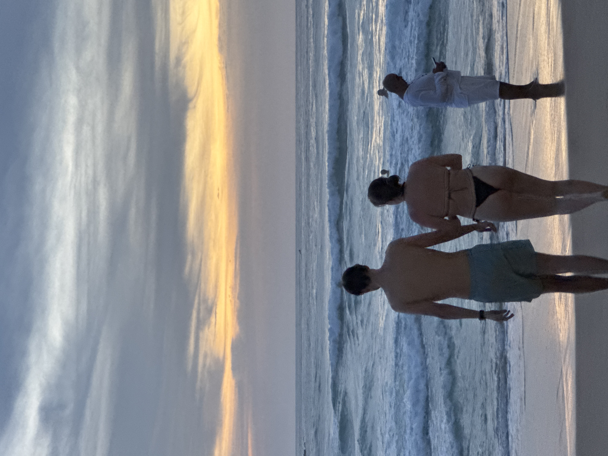 A couple walks hand-in-hand on a sandy beach at sunset in Karon, Thailand. The man wears blue and white striped swim trunks, and the woman wears a black bikini. In the background, a man in a white shirt and shorts looks at his phone, and other figures are visible in the ocean. The sky is filled with clouds painted in hues of orange and purple, reflecting on the gentle waves of the Andaman Sea.