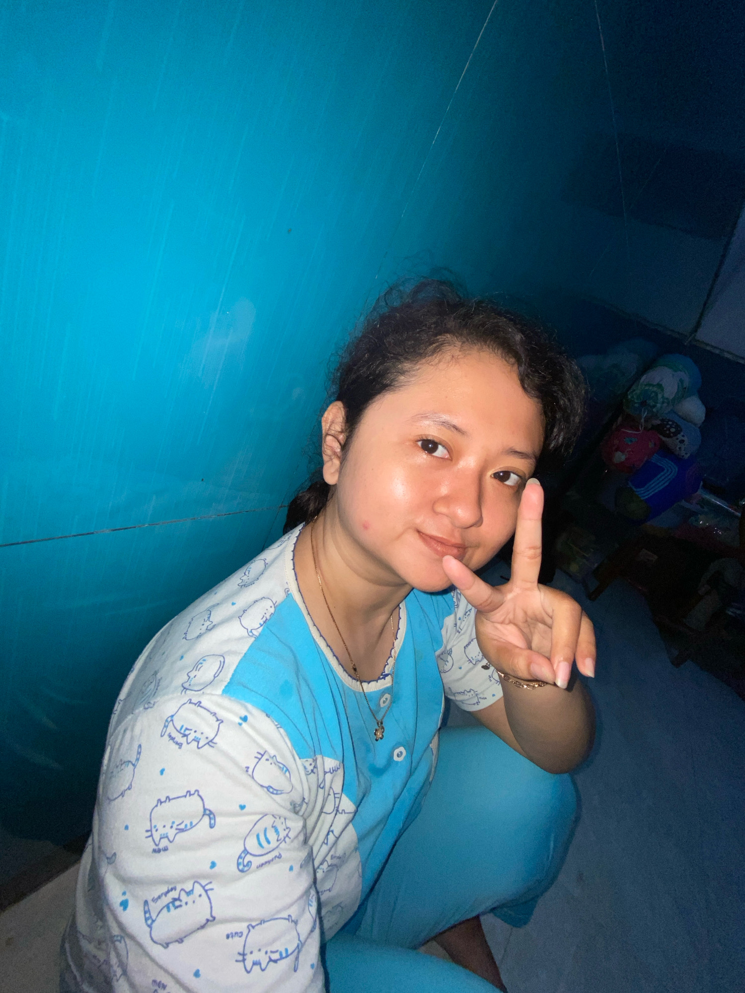 A young woman is squatting in front of a blue wall, making a peace sign with her right hand. She is wearing pajamas with a white top adorned with blue cartoon cats and solid blue pants. The room appears to be dimly lit and cluttered with various objects in the background, including what appear to be stuffed animals and storage containers. The overall impression is that of a candid, indoor photograph taken in a residential setting within the city of Gelam, Indonesia.